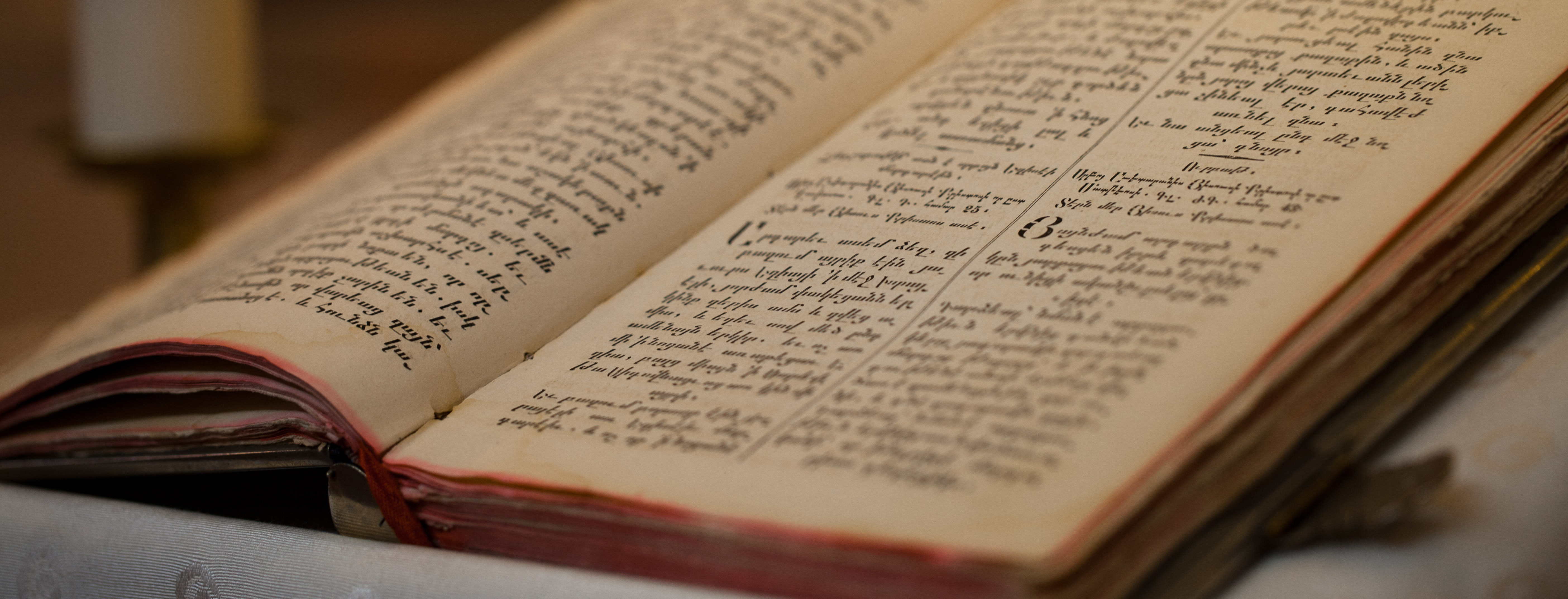 Close-up of an old open book with handwritten text in columns and a lit candle blurred in the background.