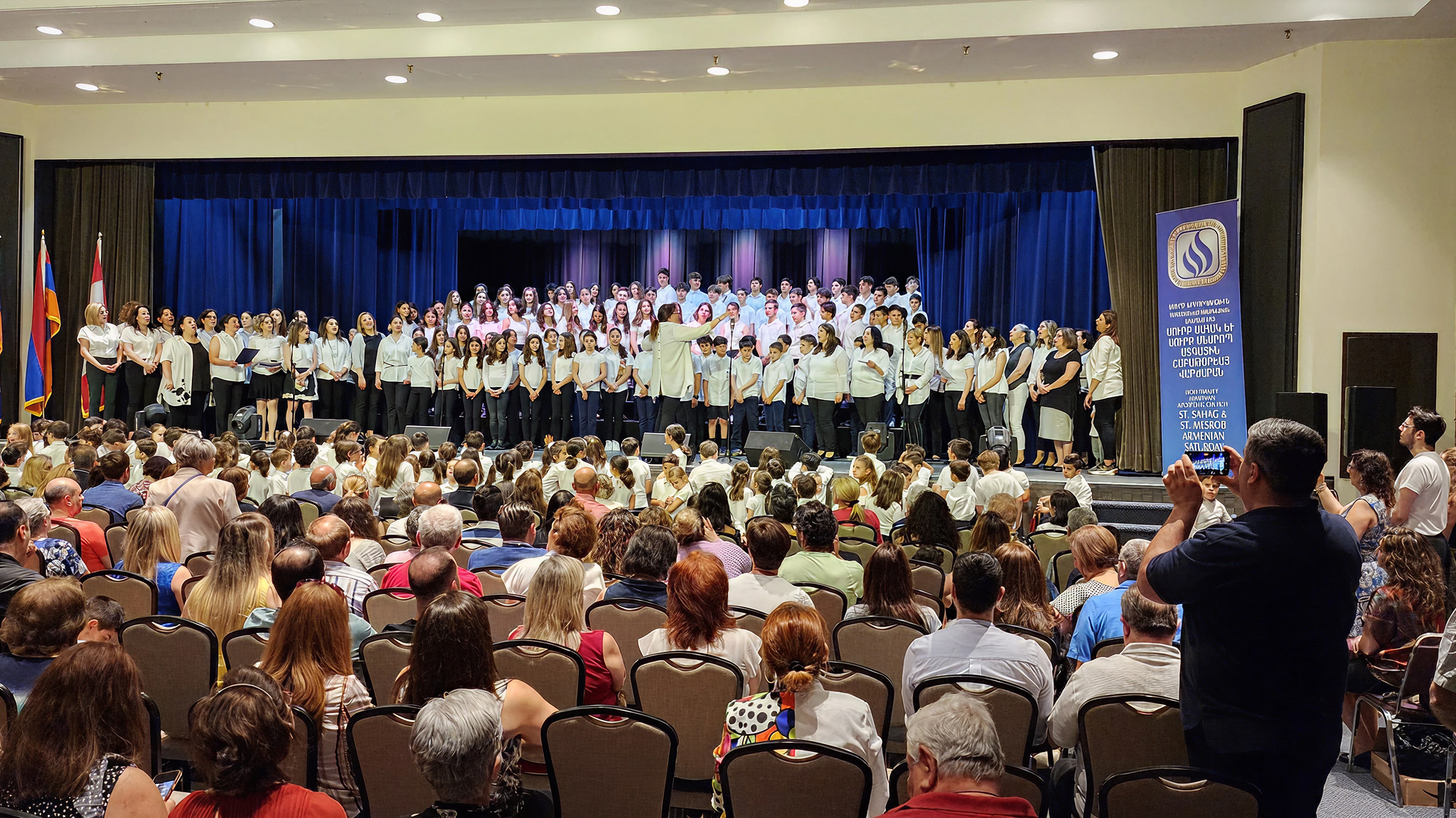 A large choir of children and adults wearing white tops perform on stage in front of an audience seated in chairs in a well-lit auditorium.