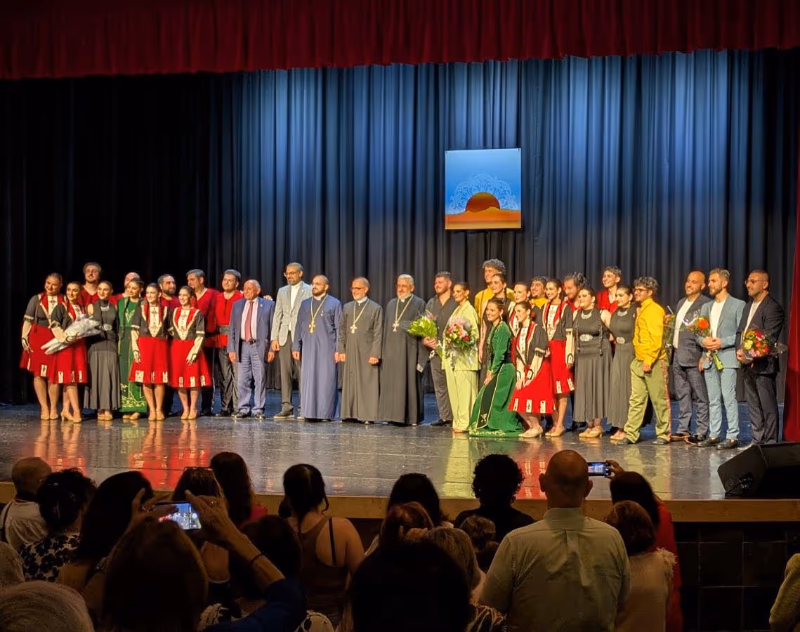 A large group of performers and clergy in traditional and formal attire pose together on a theater stage with a blue curtain and an audience in the foreground.