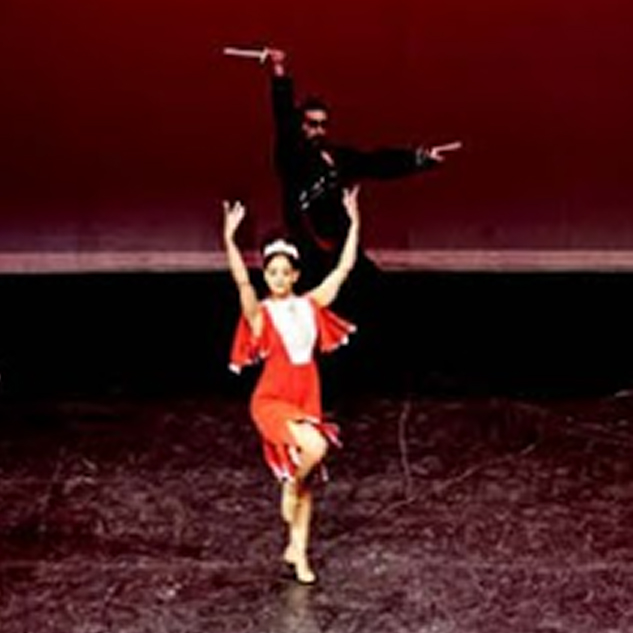 A female dancer in a red and white costume performing on stage with a male dancer in black behind her holding a sword.