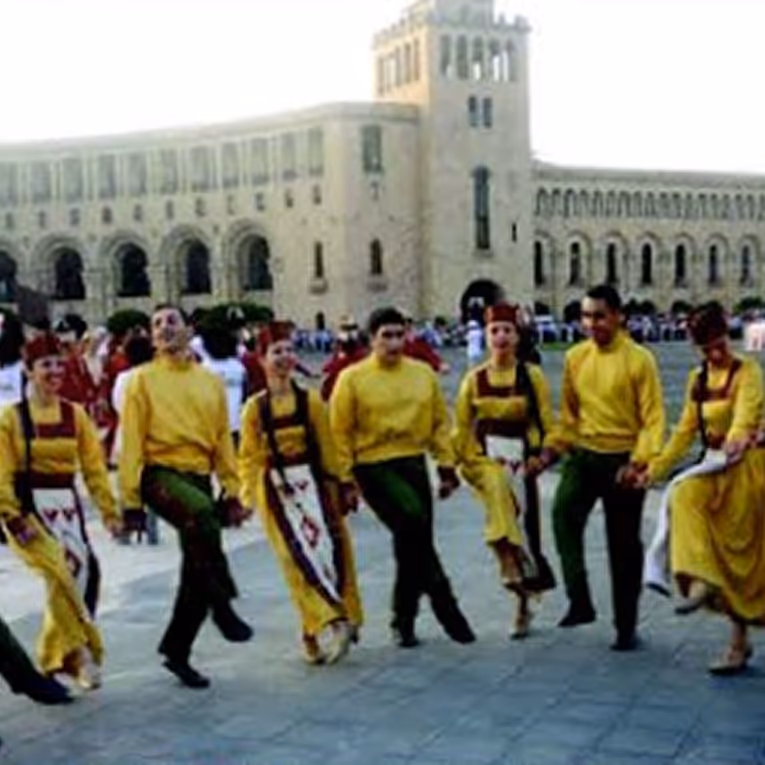 Group of dancers in traditional yellow attire performing a folk dance outdoors in front of a historic stone building.