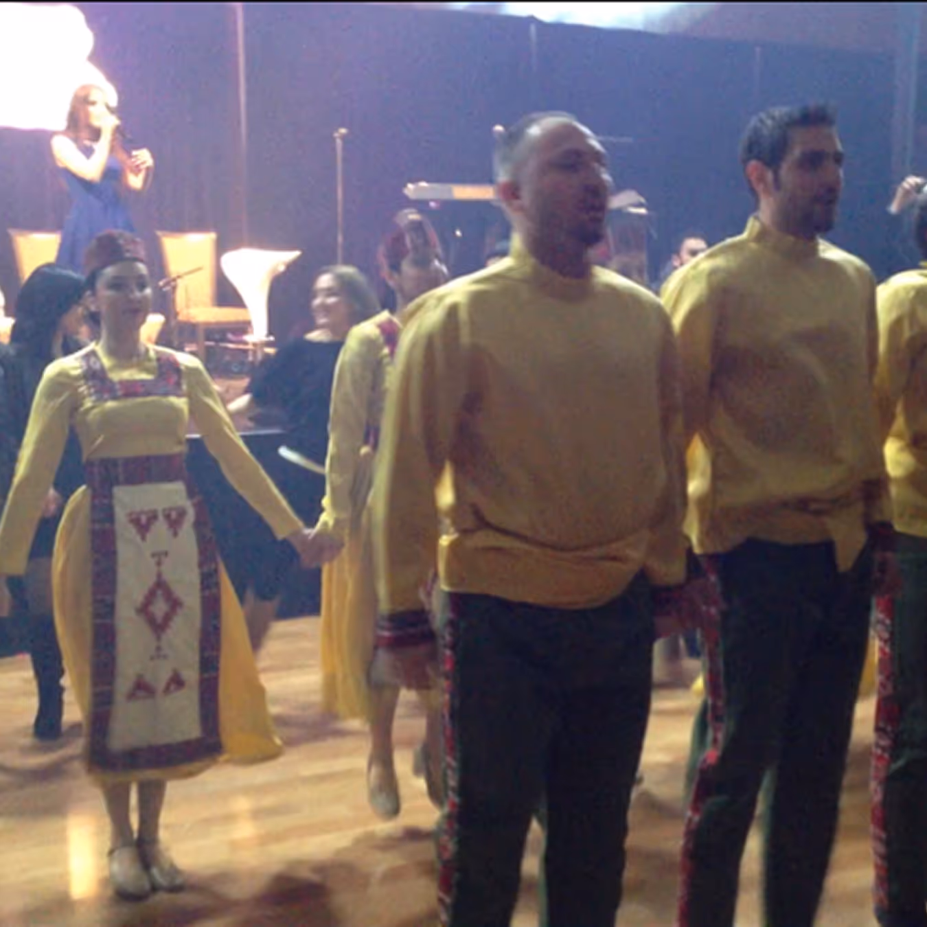 Group of people in traditional yellow and embroidered folk costumes holding hands and dancing on a wooden floor with a singer performing on stage in the background.
