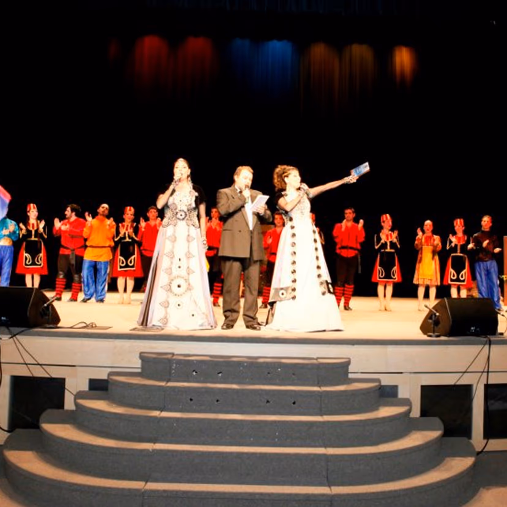 Three presenters on stage with microphones in front of a group of performers in colorful traditional costumes, with a curved staircase leading to the stage.