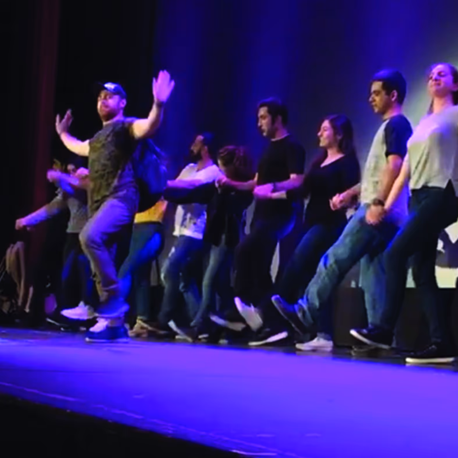 A group of people performing a coordinated dance on stage under purple lighting.