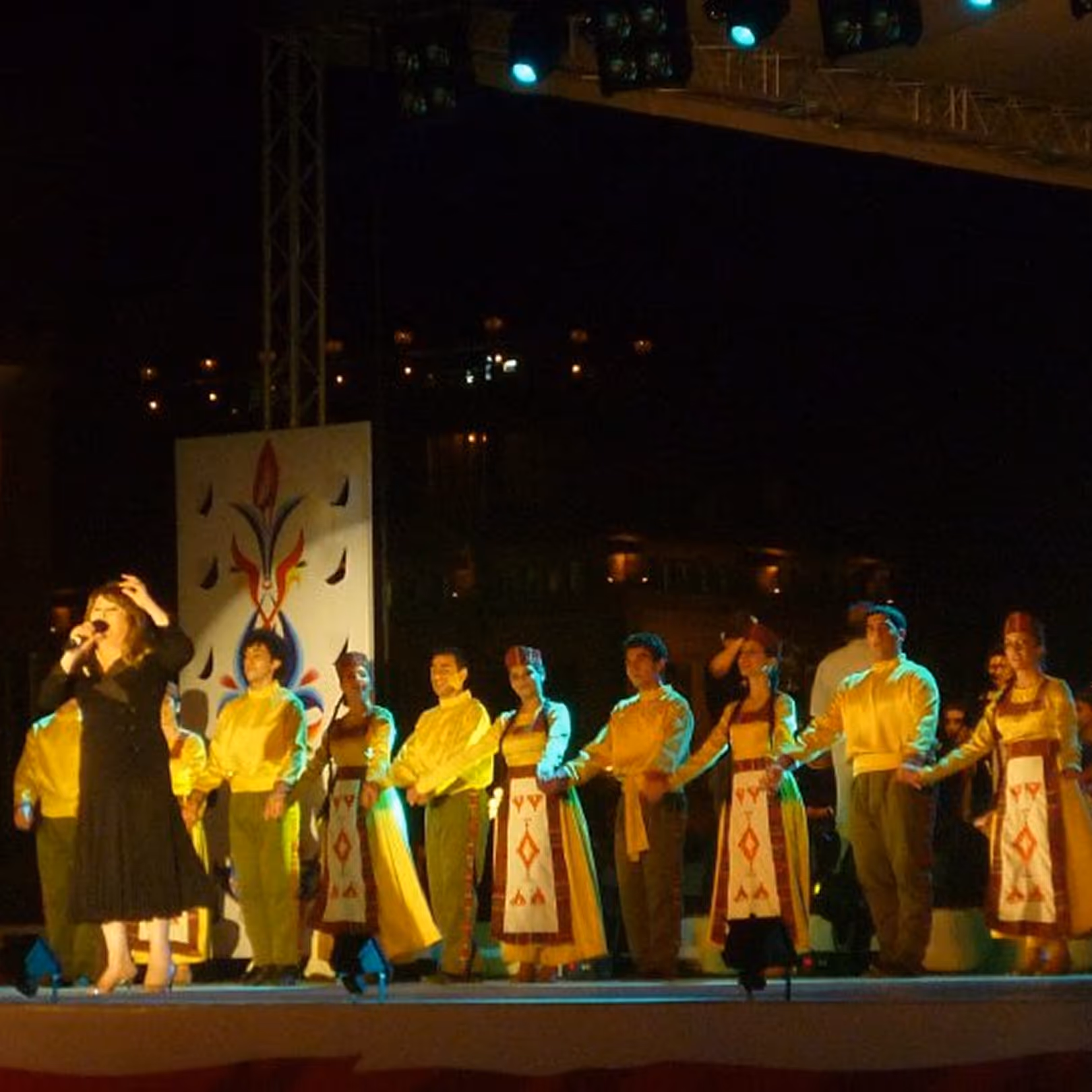 Group of performers on stage in traditional yellow and red costumes with a female singer on the left holding a microphone.