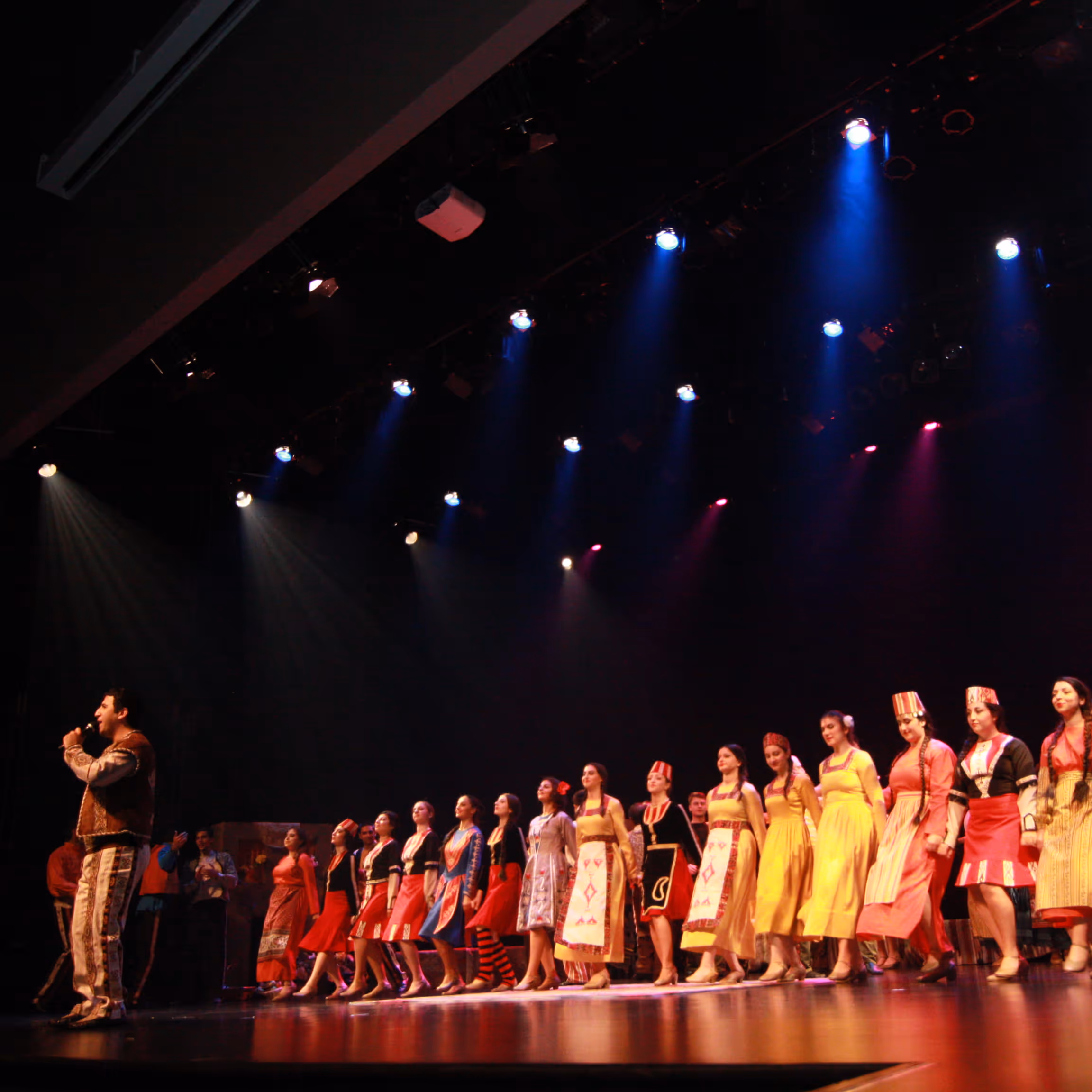Group of performers in colorful traditional costumes lined up on stage under spotlights with a male singer holding a microphone on the left.