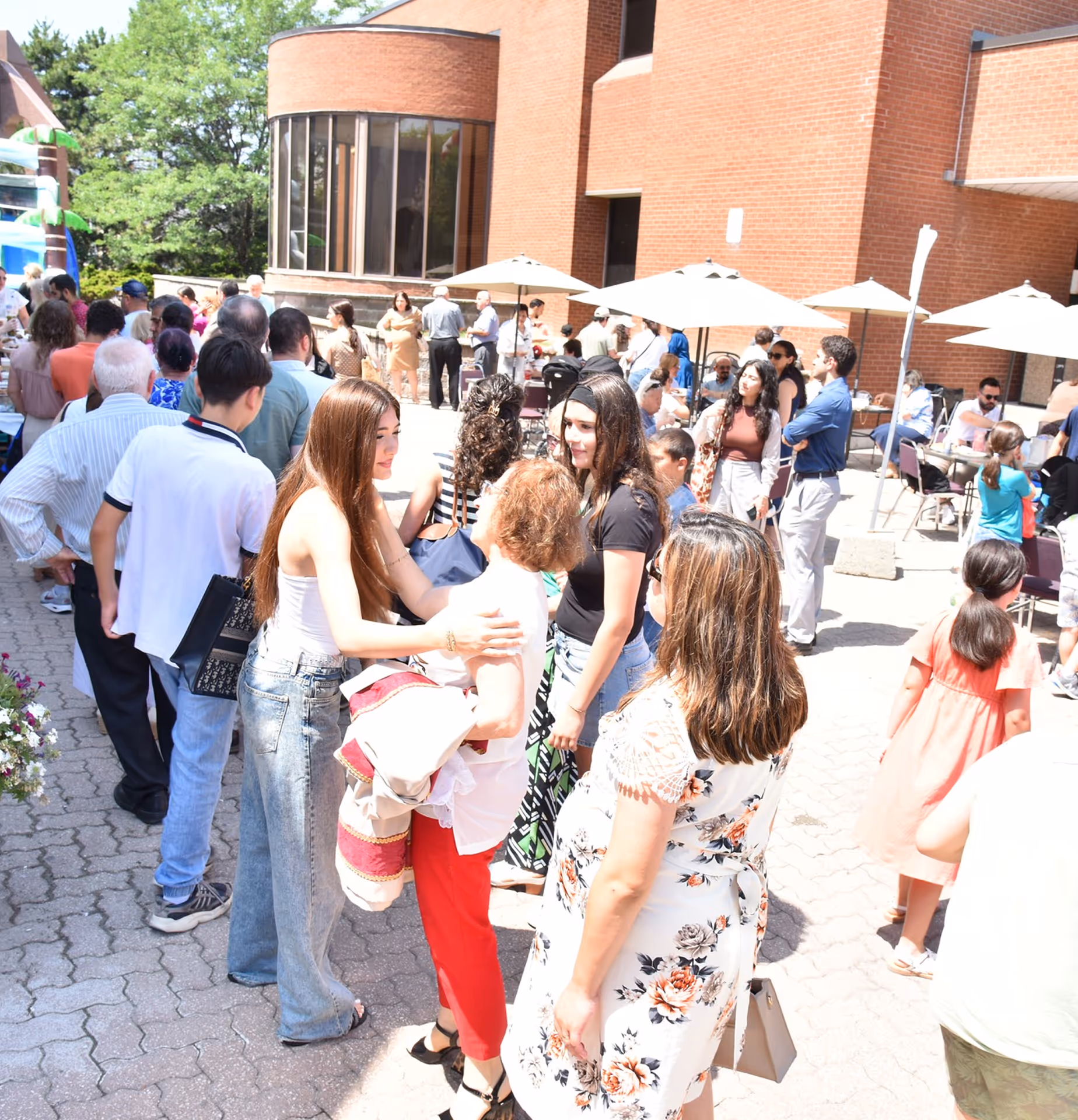 A group of people socializing outdoors on a sunny day near a brick building with tables and umbrellas.
