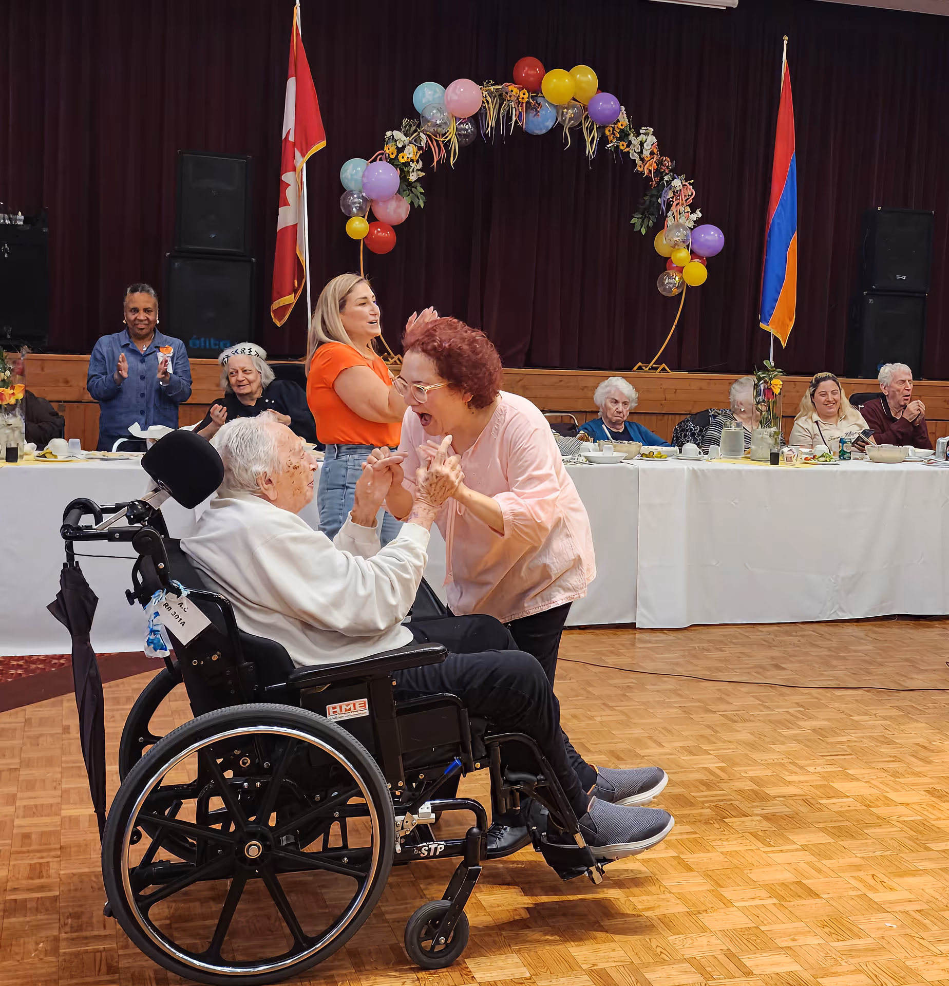 An elderly man in a wheelchair joyfully holding hands and interacting with a woman in a pink shirt at a celebratory event with seated guests and colorful balloons.