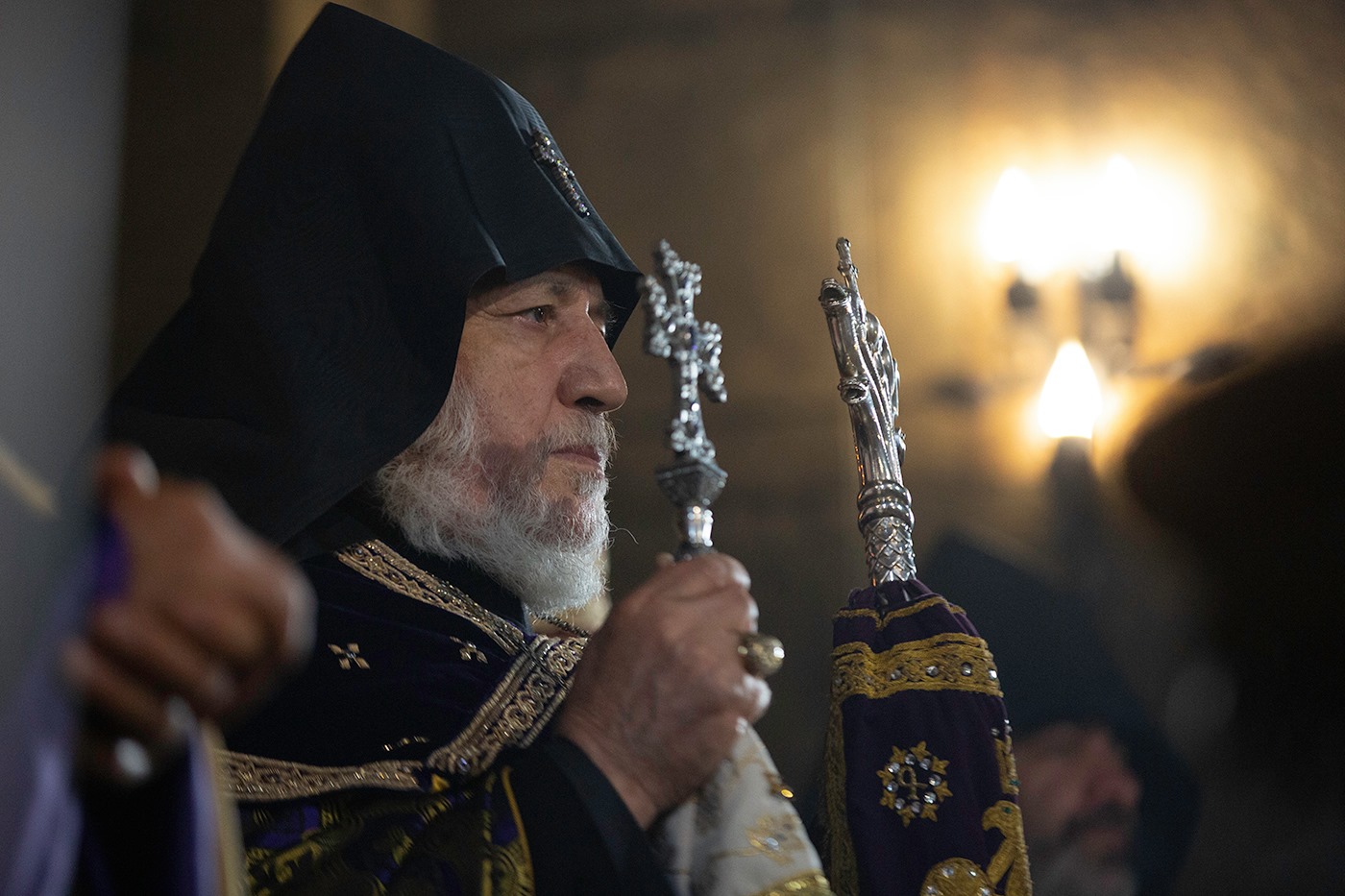 Elderly clergyman with a white beard wearing a black hooded religious robe holding ornate silver religious staffs.