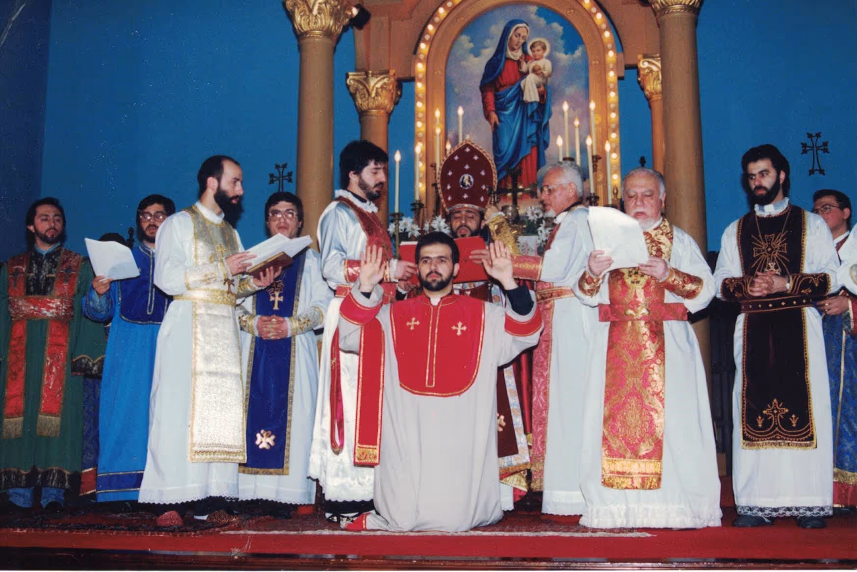 A group of priests in liturgical vestments conducting a religious ceremony inside a church with a painting of Madonna and child in the background.