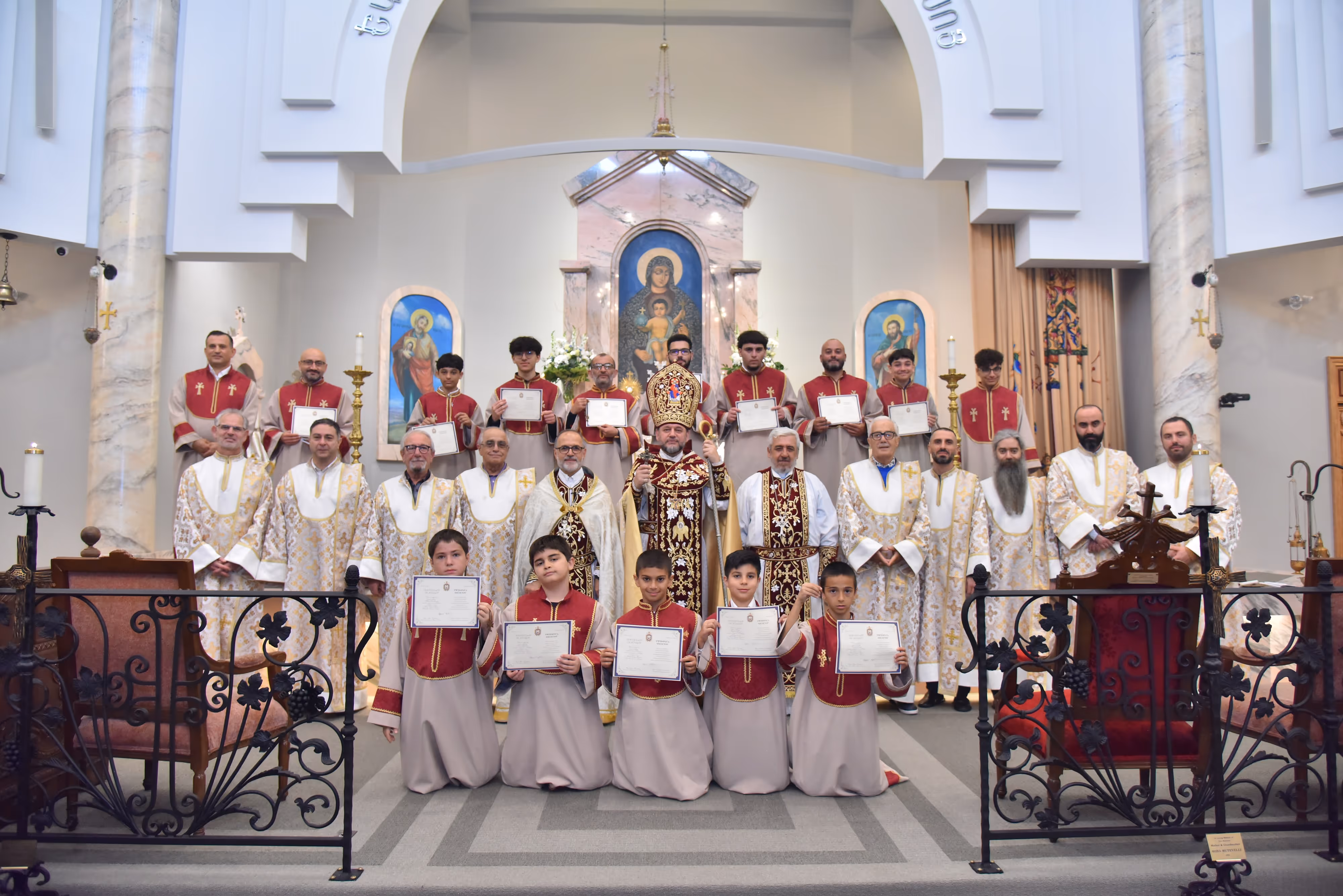 Group of clergy and altar boys in church holding certificates during a religious ceremony under arches and religious icons.