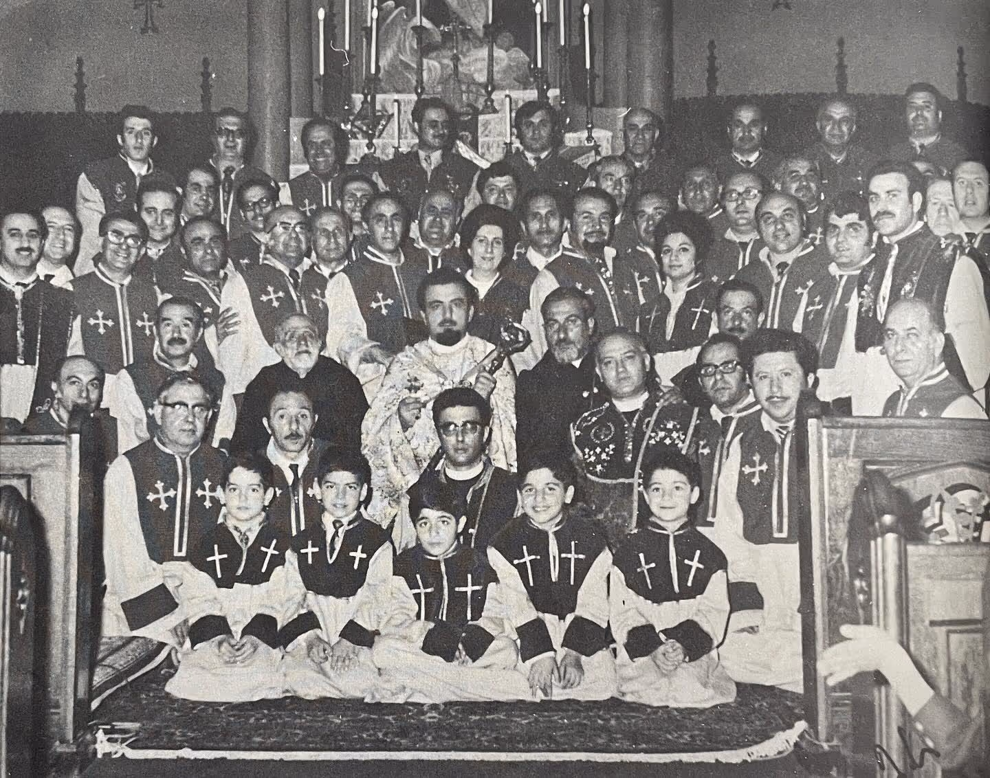 Black and white group photo of approximately 50 people, including men, women, and boys, many wearing ceremonial or religious robes with crosses, gathered inside a church in front of an altar.