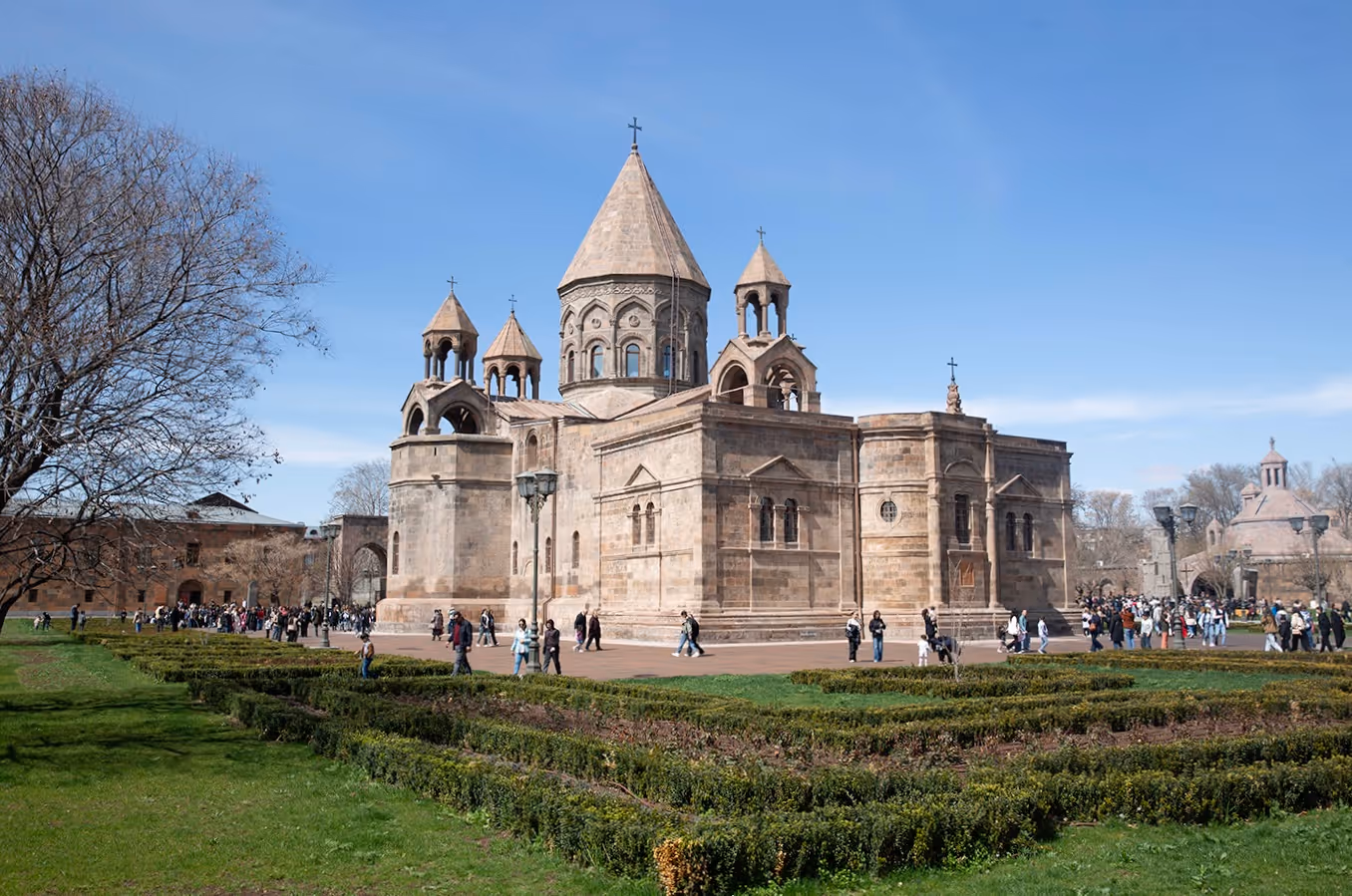 Medieval stone church with conical towers and crosses surrounded by people and landscaped gardens under a clear blue sky.