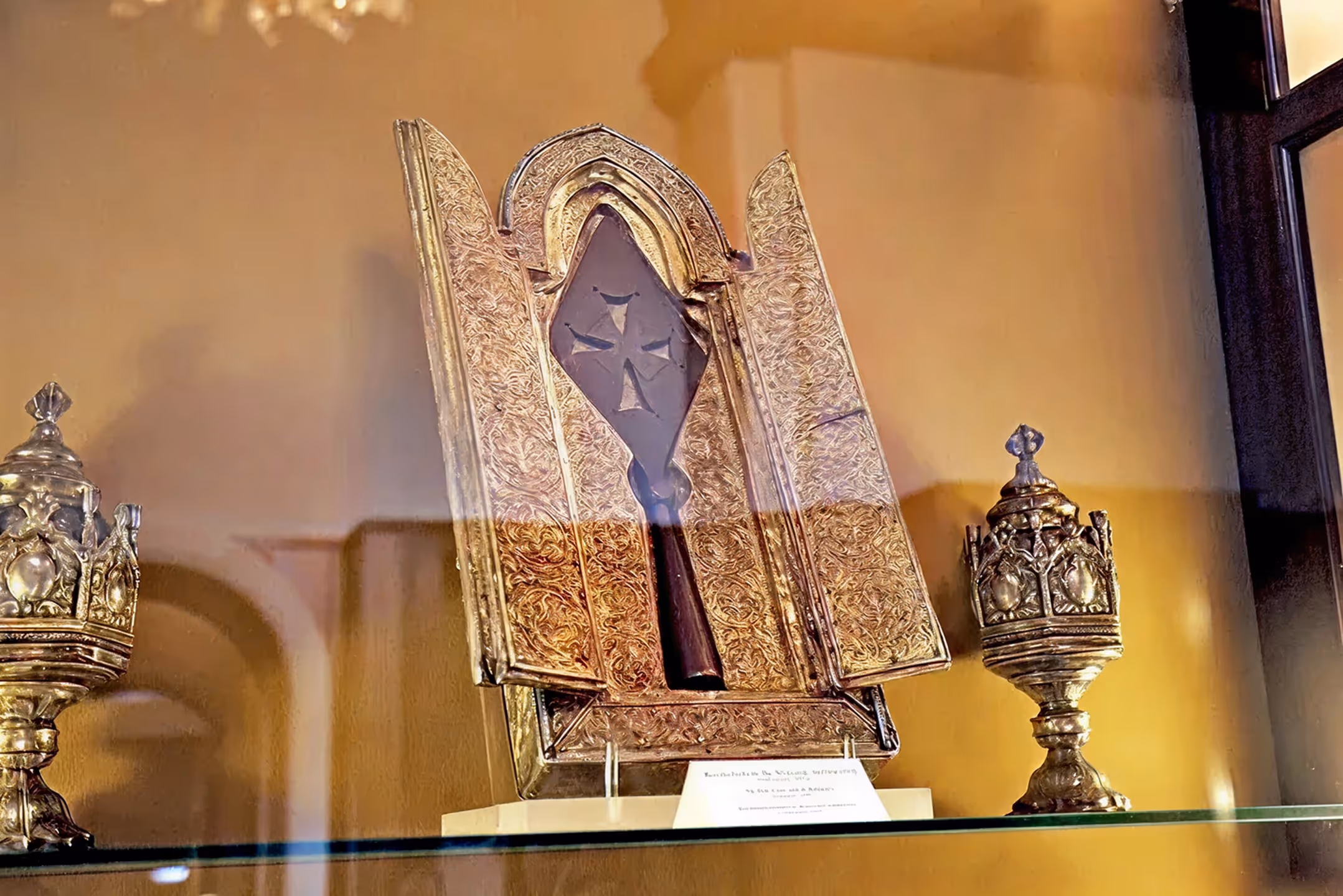 Ornate religious relics displayed in a glass case, including a decorated gold-covered object with a cross and two intricate chalices.