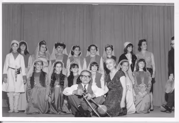 Black and white photo of a group of children and two adults, some in traditional costumes, posing on stage with a curtain backdrop; one adult is seated holding a musical instrument.