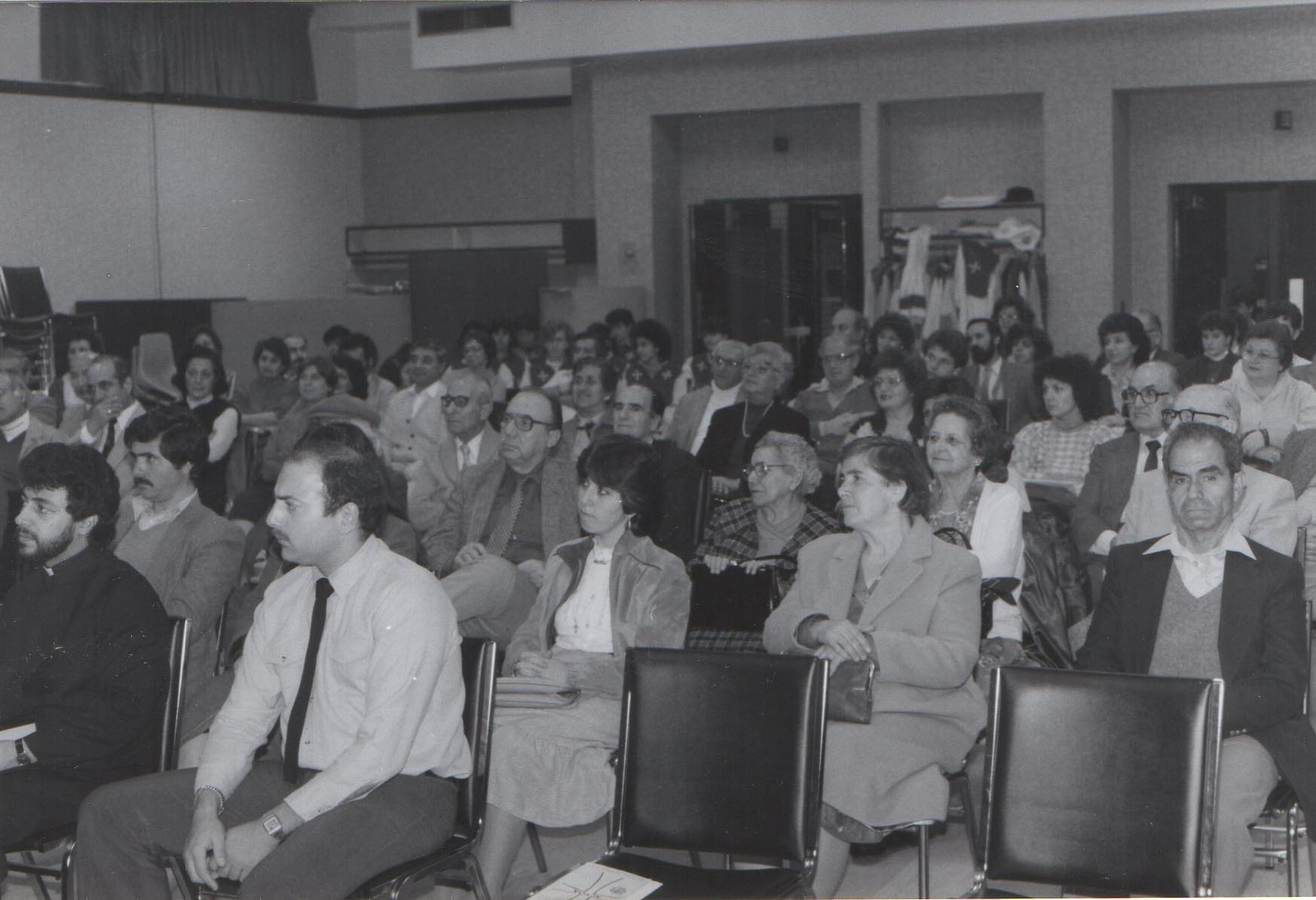 Black and white photo of a mixed-age audience seated in rows, attentively watching something out of frame in an indoor setting.
