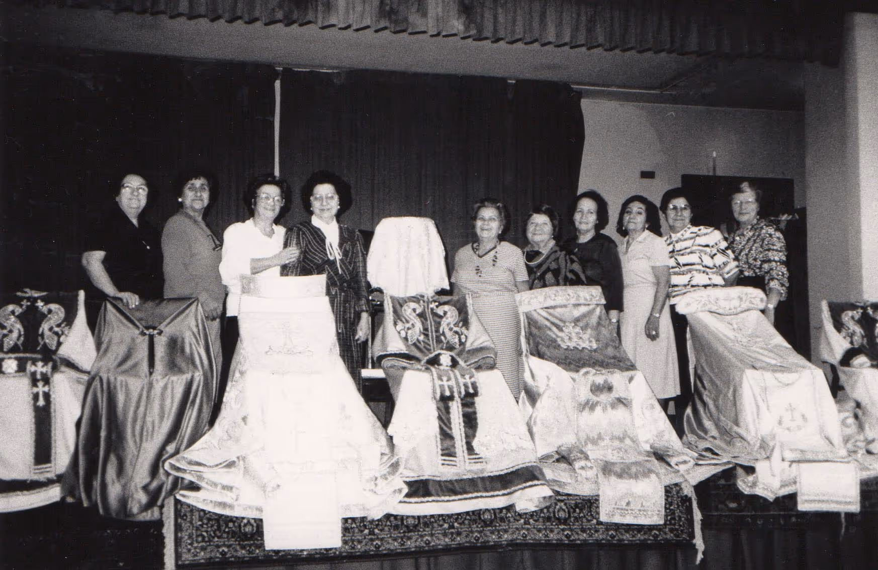 Black and white photo of ten women standing behind a table displaying multiple intricately embroidered religious vestments.