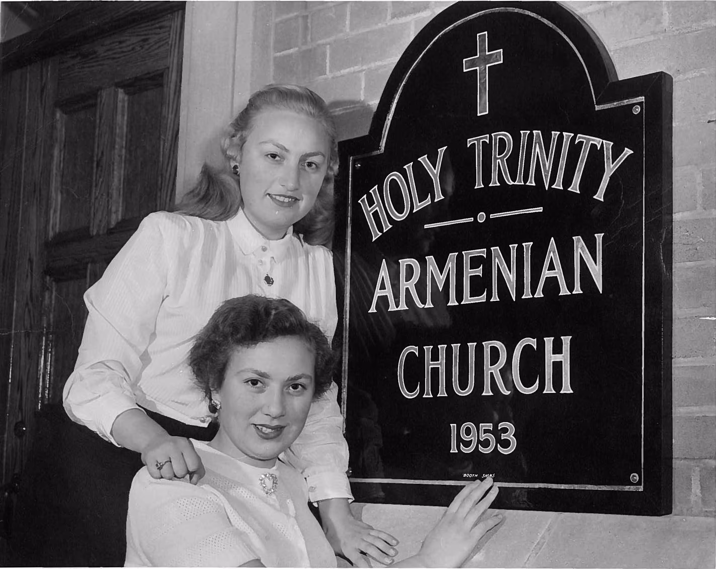 Two women posing next to a sign reading 'Holy Trinity Armenian Church 1953' mounted on a brick wall.