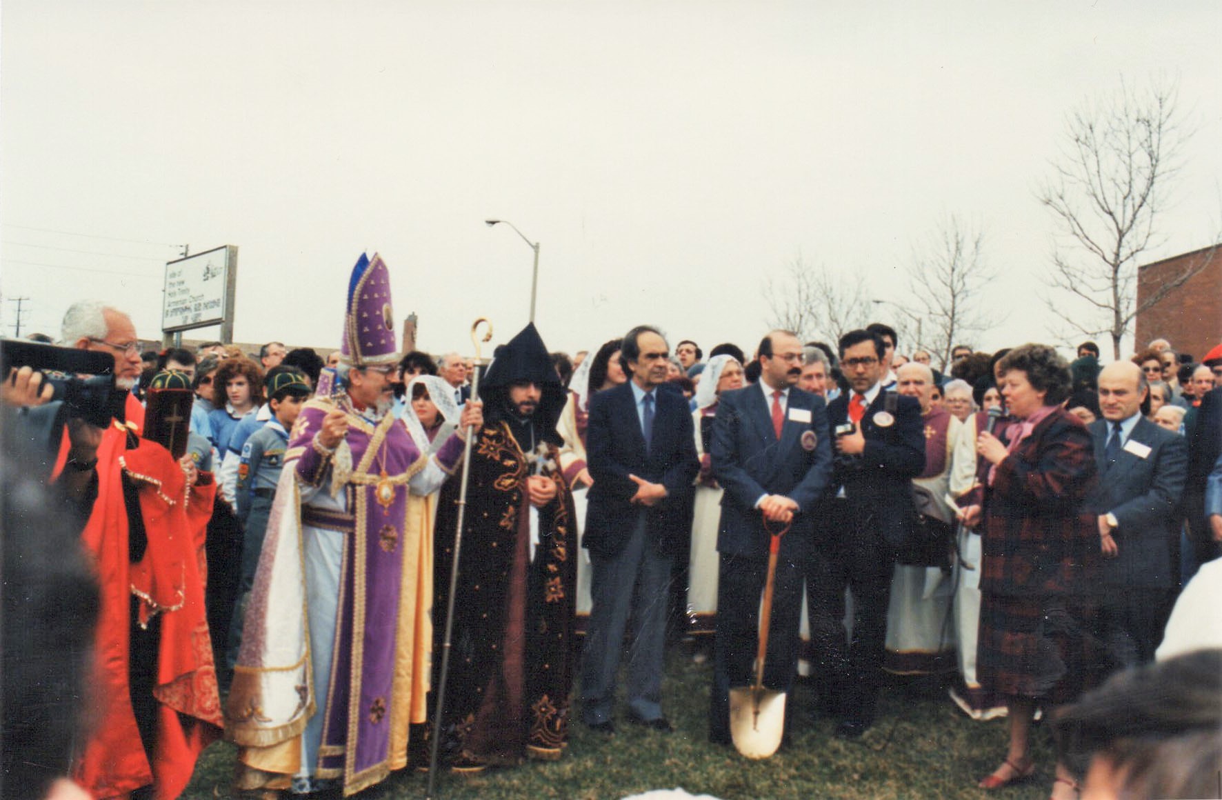 Crowd gathered outdoors for a ceremonial event with religious leaders in ornate robes and a man holding a shovel on grass.