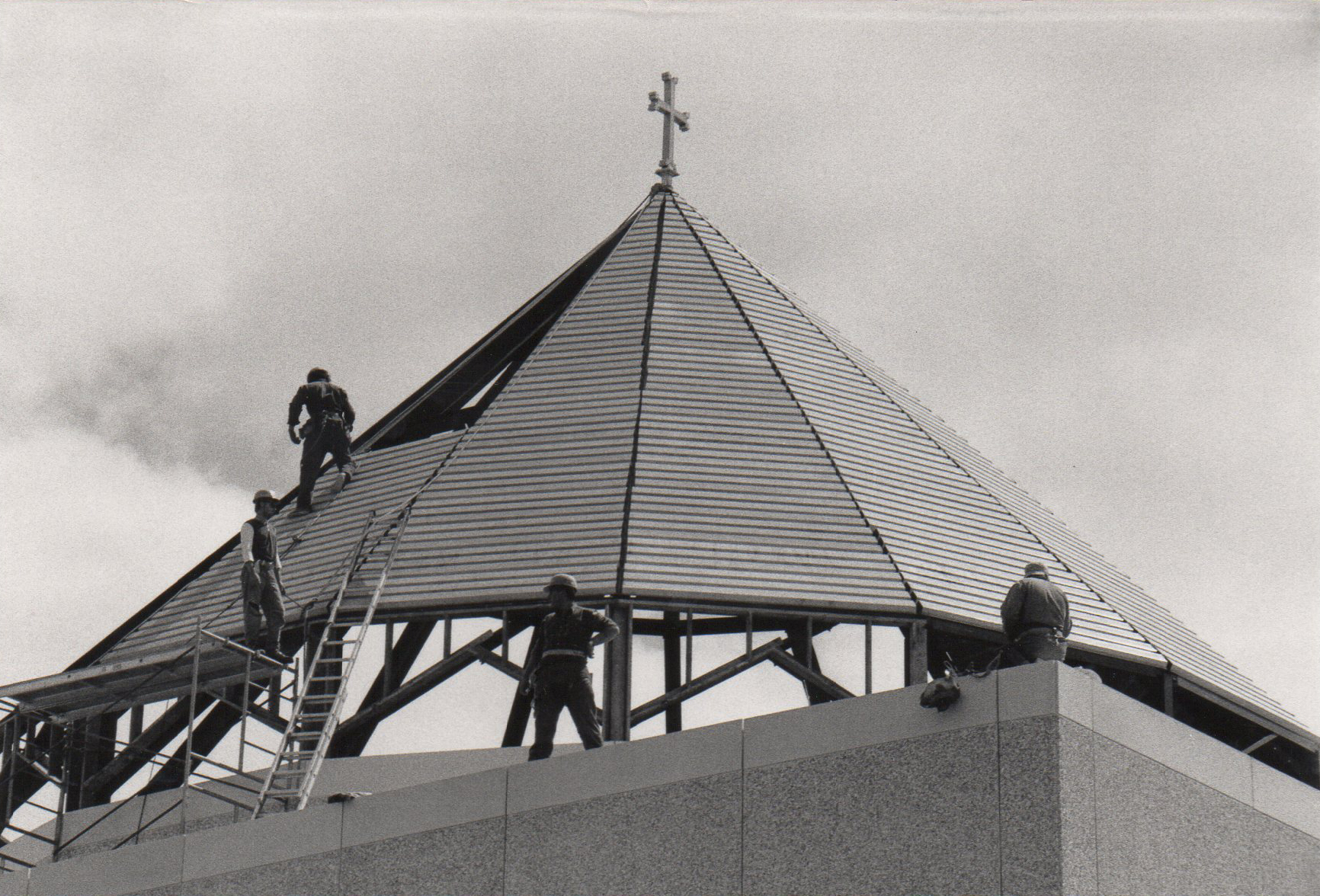Four construction workers wearing helmets working on the roof of a building with a large cross at the top.