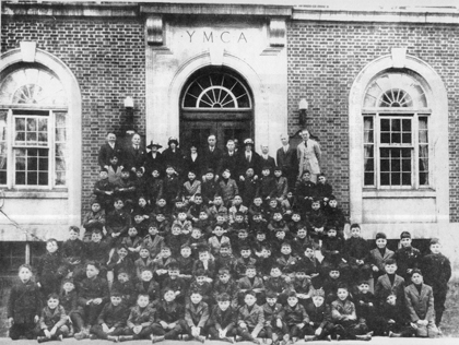 Black and white photo of a large group of children and adults posing in front of a YMCA brick building with an arched entrance.