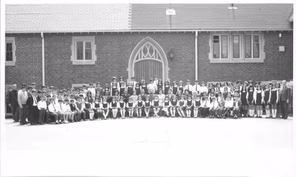 Black and white group photo of a large group of school children and adults posing outside a brick building with a large arched wooden door.
