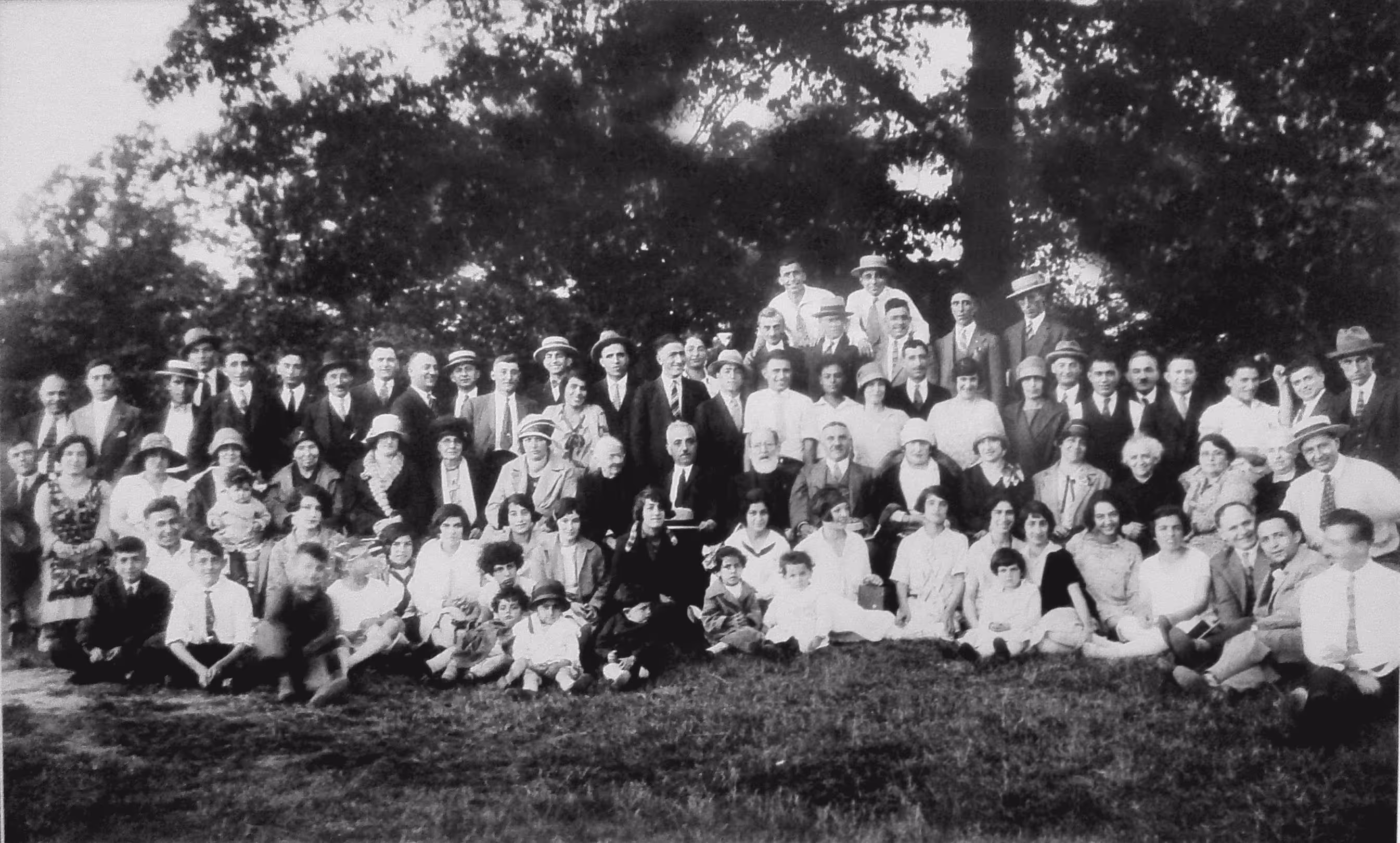 Large black and white group photo of men, women, and children dressed in early 20th-century attire, seated and standing outdoors on grass with trees in the background.