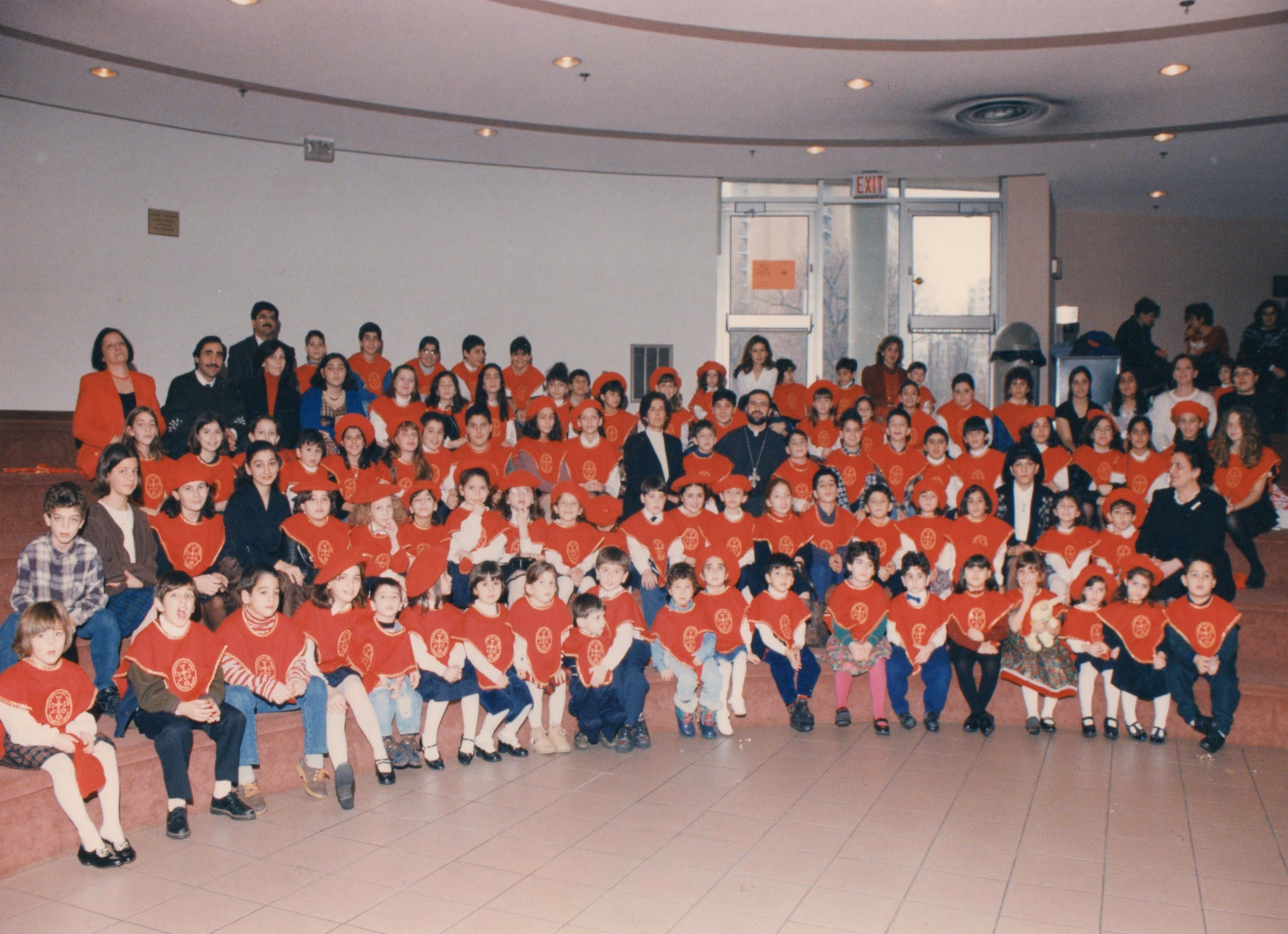 Large group of children and adults posing indoors on steps, many children wearing red uniforms with matching berets.