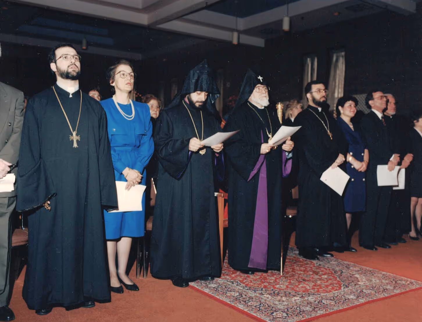 Group of clergy and attendees standing in a formal setting, some holding papers, with religious robes and crosses visible.