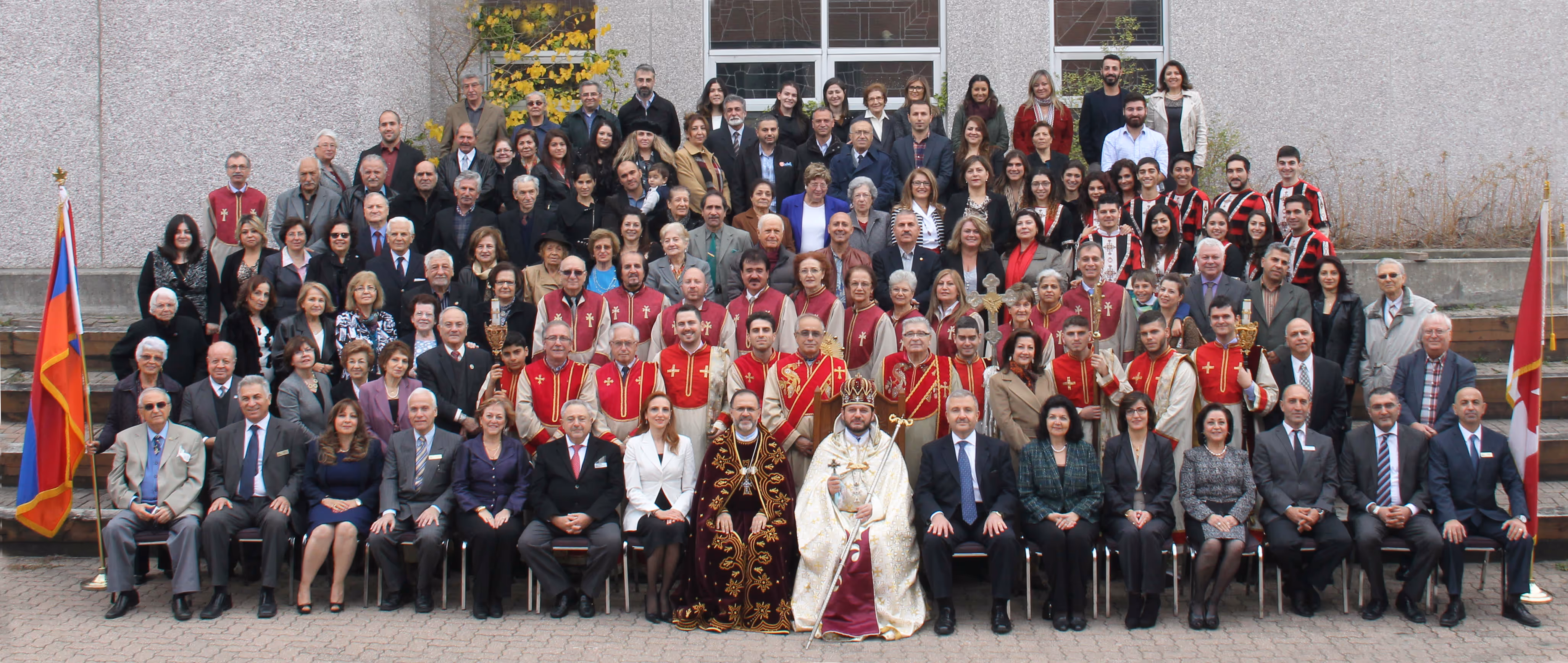 Large group portrait of men and women, including clergy in ornate robes, standing and sitting outdoors with two flags on either side.