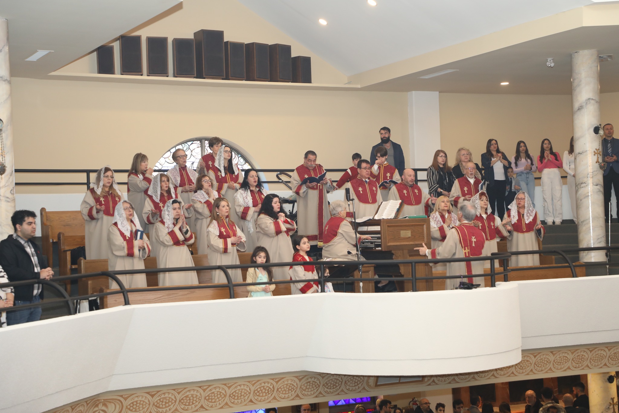 Church choir dressed in beige and red robes singing on a balcony with a conductor and a small organist, with some audience members standing behind them.