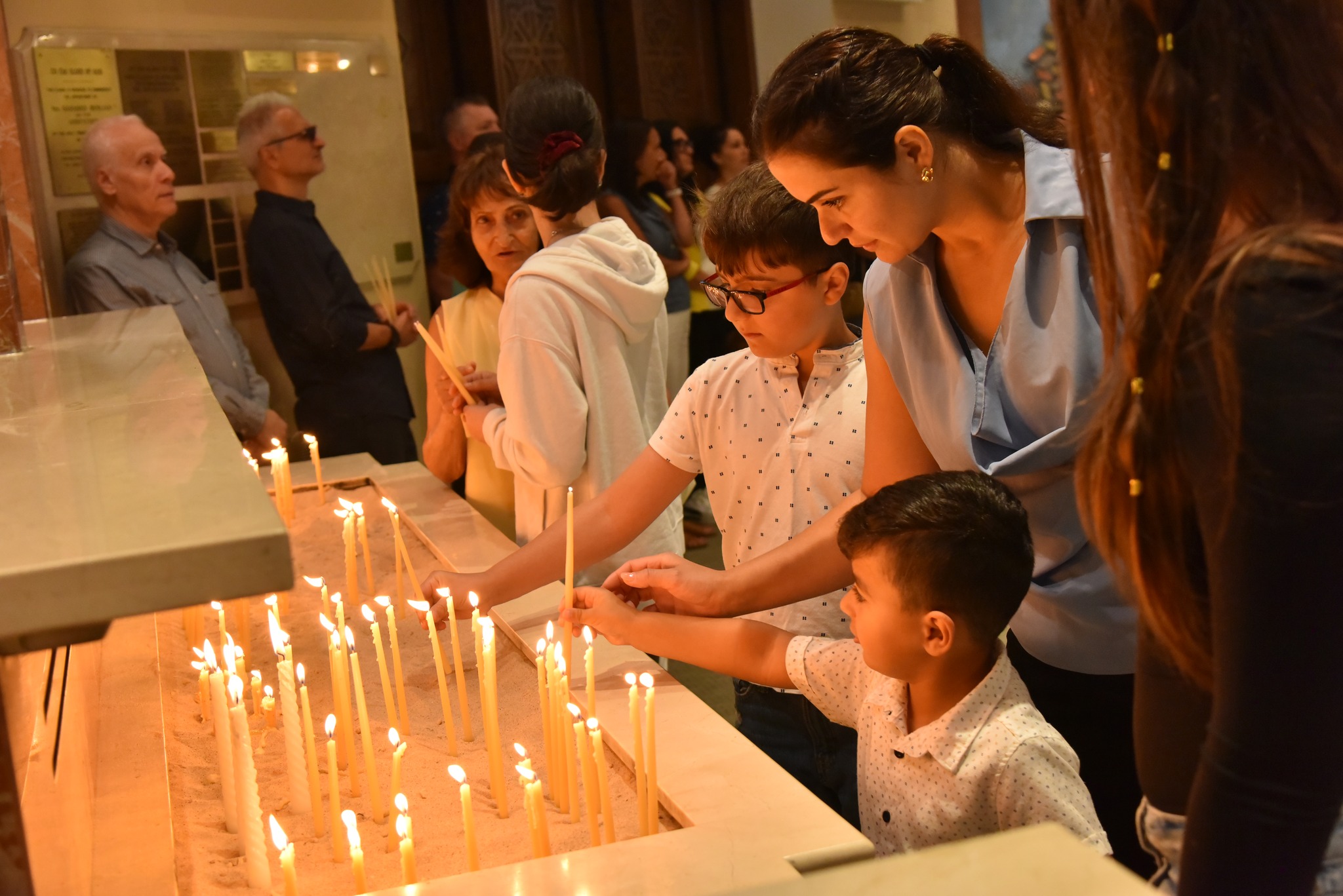 People lighting thin candles in a candle rack inside a church or chapel.
