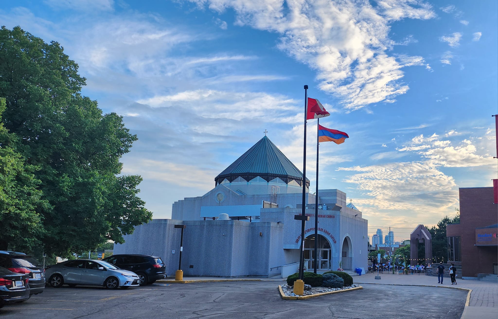 View of a church building with a conical roof, flags on poles, parked cars, and people walking near an outdoor seating area under a partly cloudy sky.