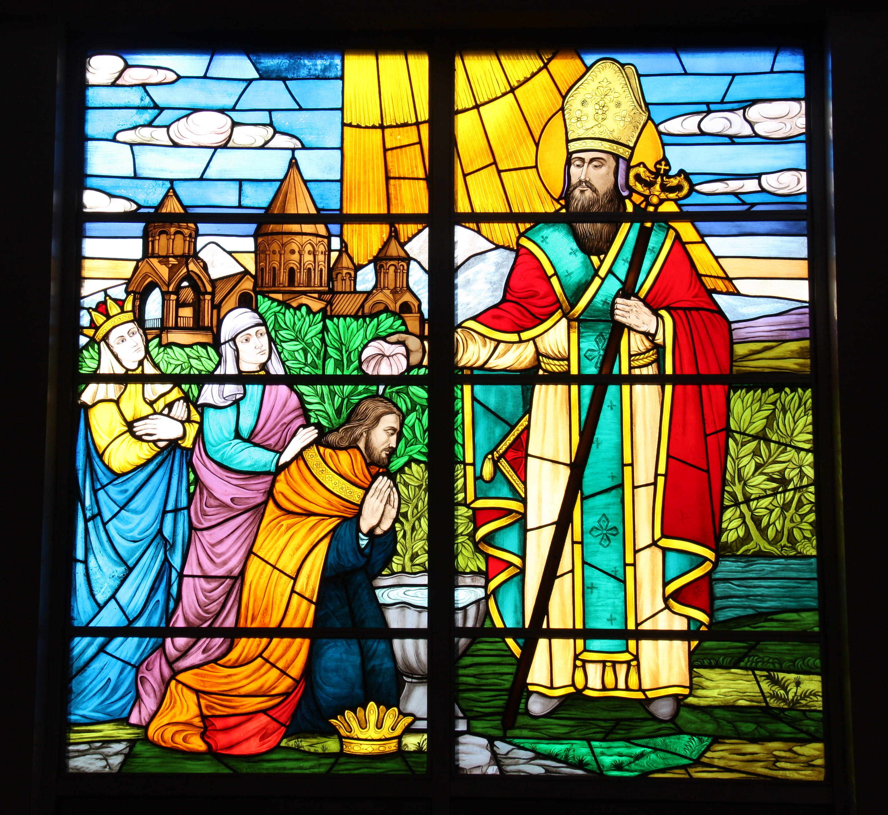 Stained glass window depicting a religious scene with a bishop blessing a kneeling man, two women standing behind, and a backdrop of churches and landscape.