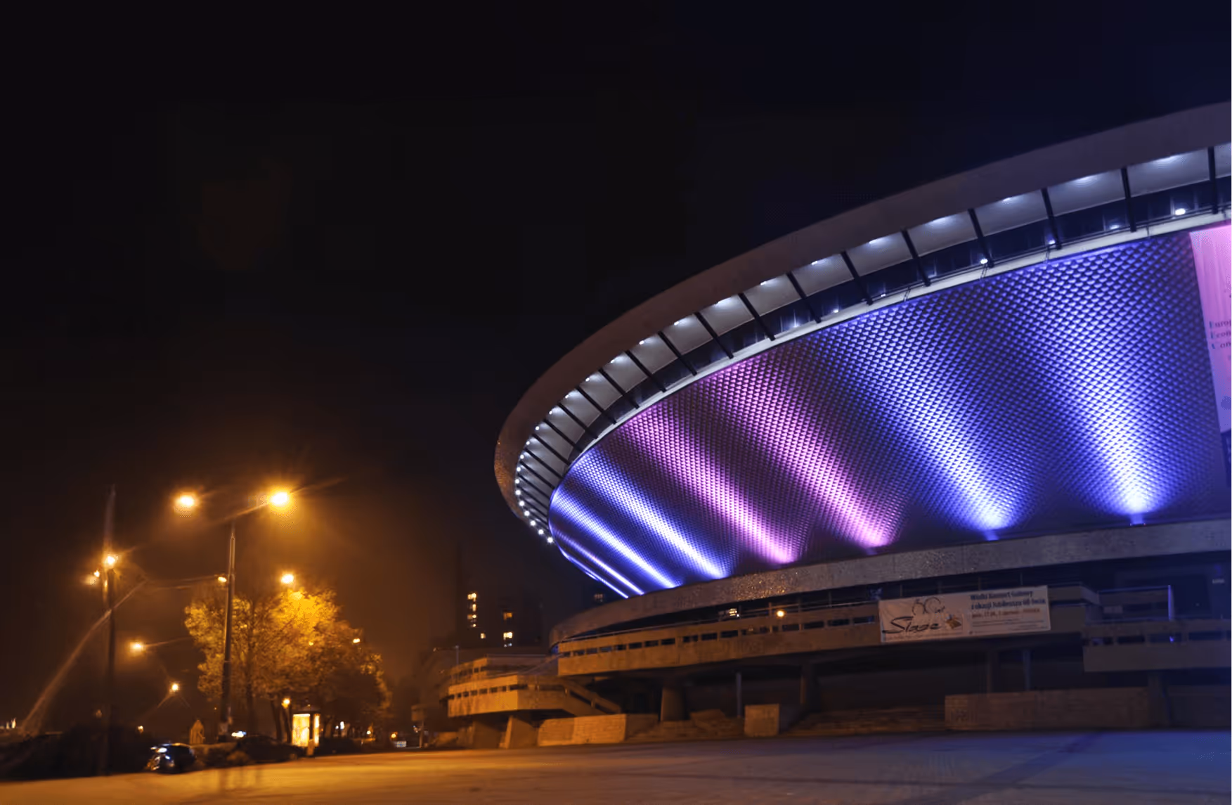 Modern circular building illuminated with purple and blue lights at night, with street lamps lighting nearby trees and a parked car.