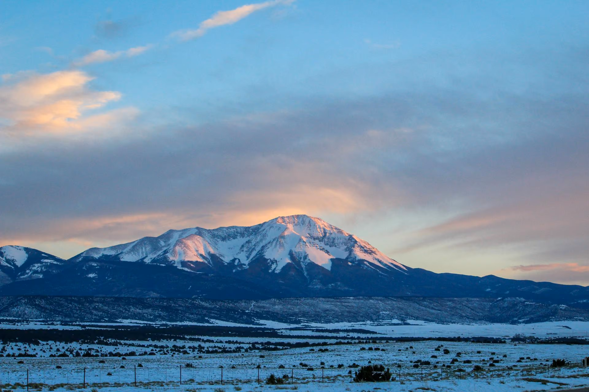 Snow-capped mountain peak at sunset with a snowy field and scattered bushes in foreground.