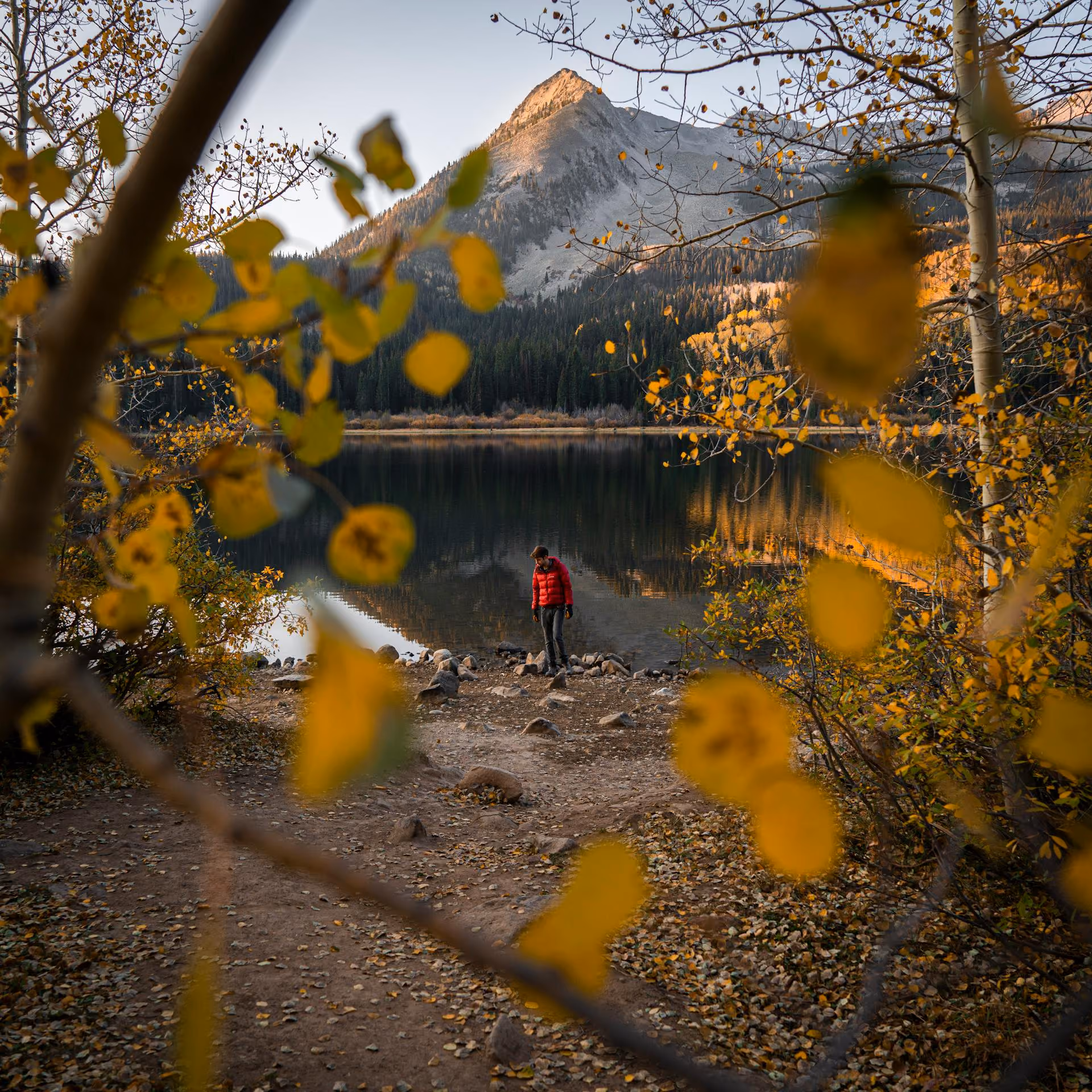 Person in a red jacket standing by a calm lake surrounded by autumn foliage and mountains.