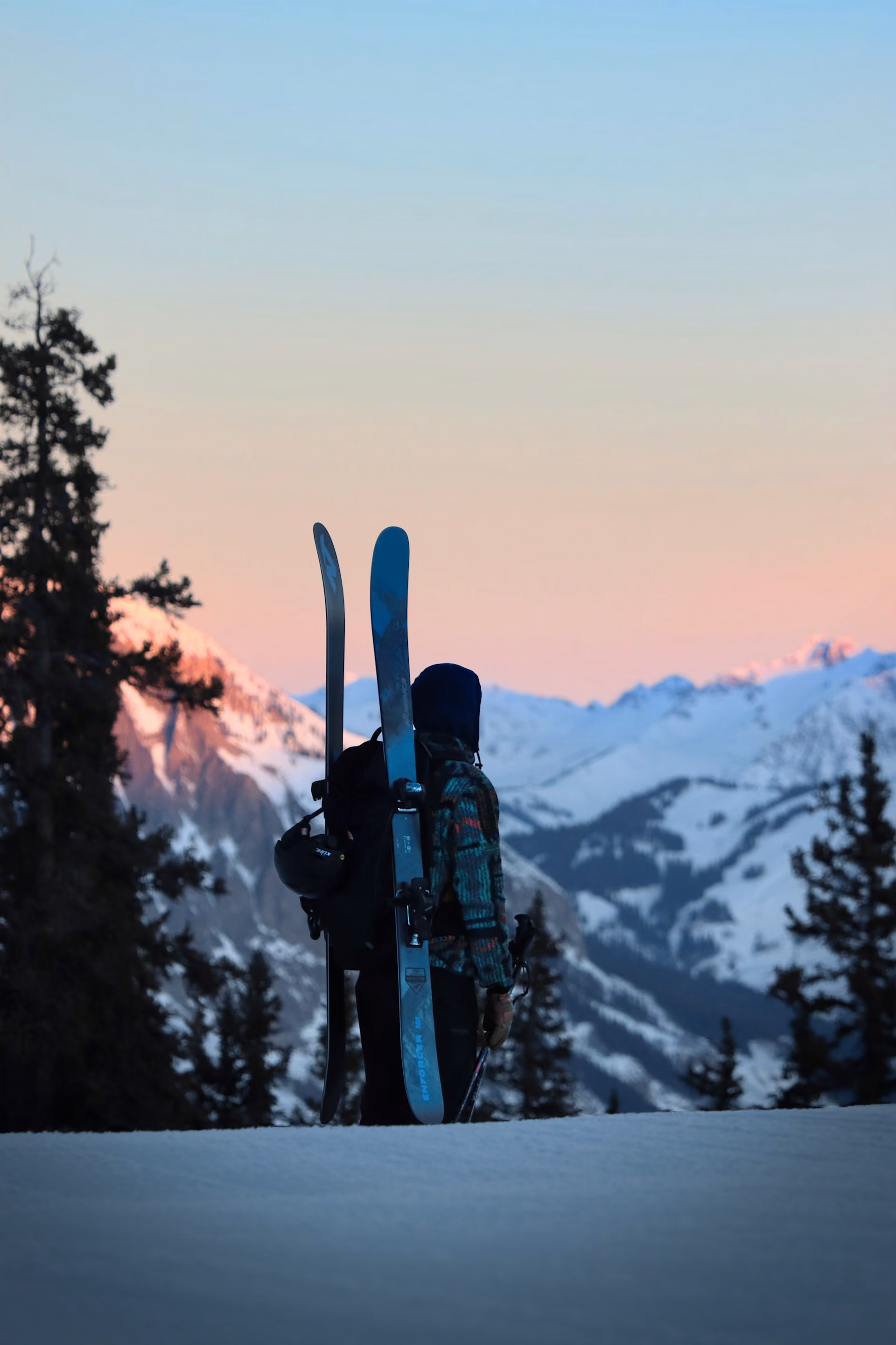 Person carrying skis and poles stands on snow looking at snowy mountain peaks at sunset.