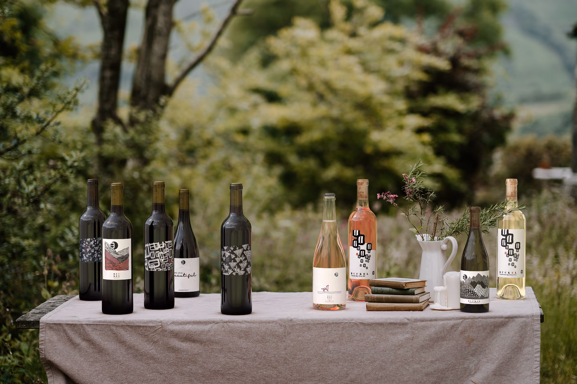 Nine wine bottles on a tablecloth-covered table outdoors with books and a white pitcher holding flowers.