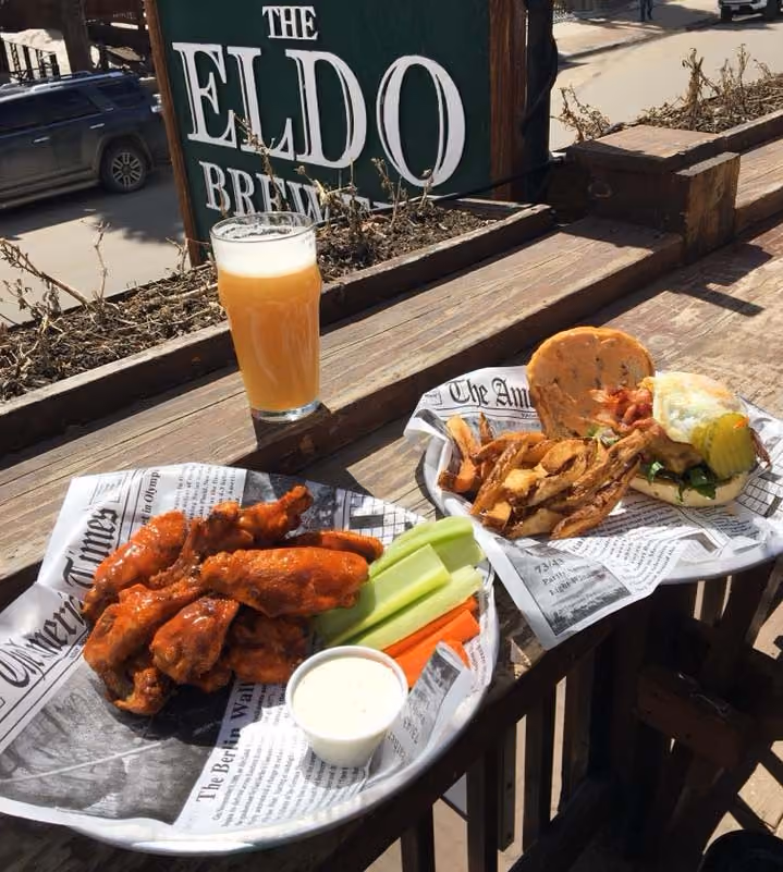 Buffalo wings with celery, carrots, ranch, burger with fries, and a glass of beer on wooden railing.