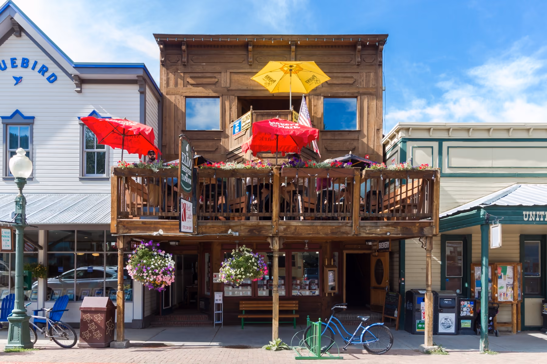 Wooden two-story building with deck seating, colorful umbrellas, flowers, and a parked blue bicycle.
