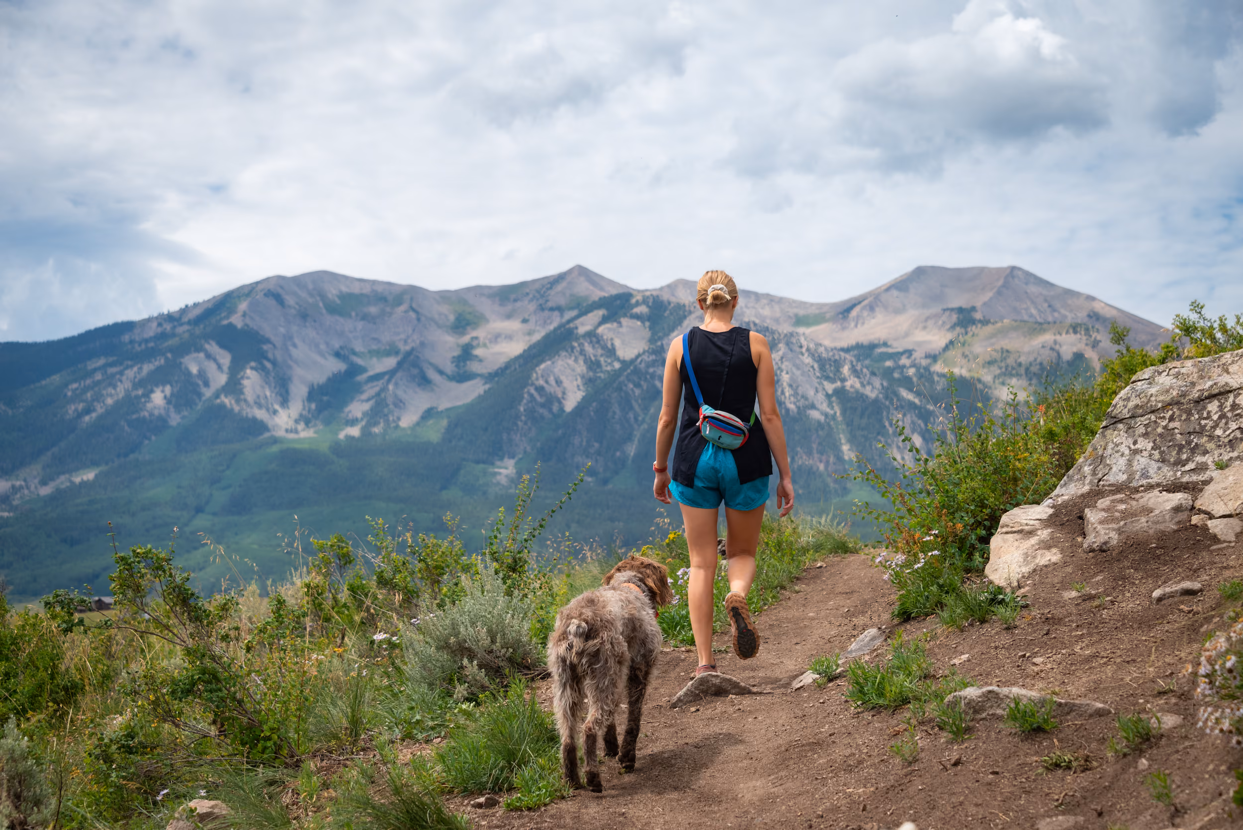 Woman hiking on a dirt trail with a dog, mountains and cloudy sky in the background.