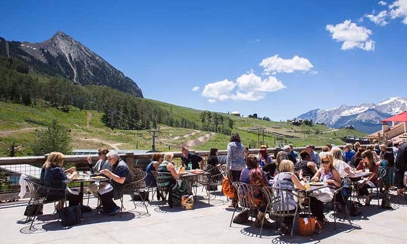 People dining outdoors on a sunny patio with mountain and green hills in the background under a blue sky.