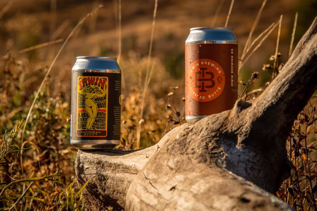 Two Irwin Brewing Co beer cans placed on a log in a sunlit grassy outdoor setting.