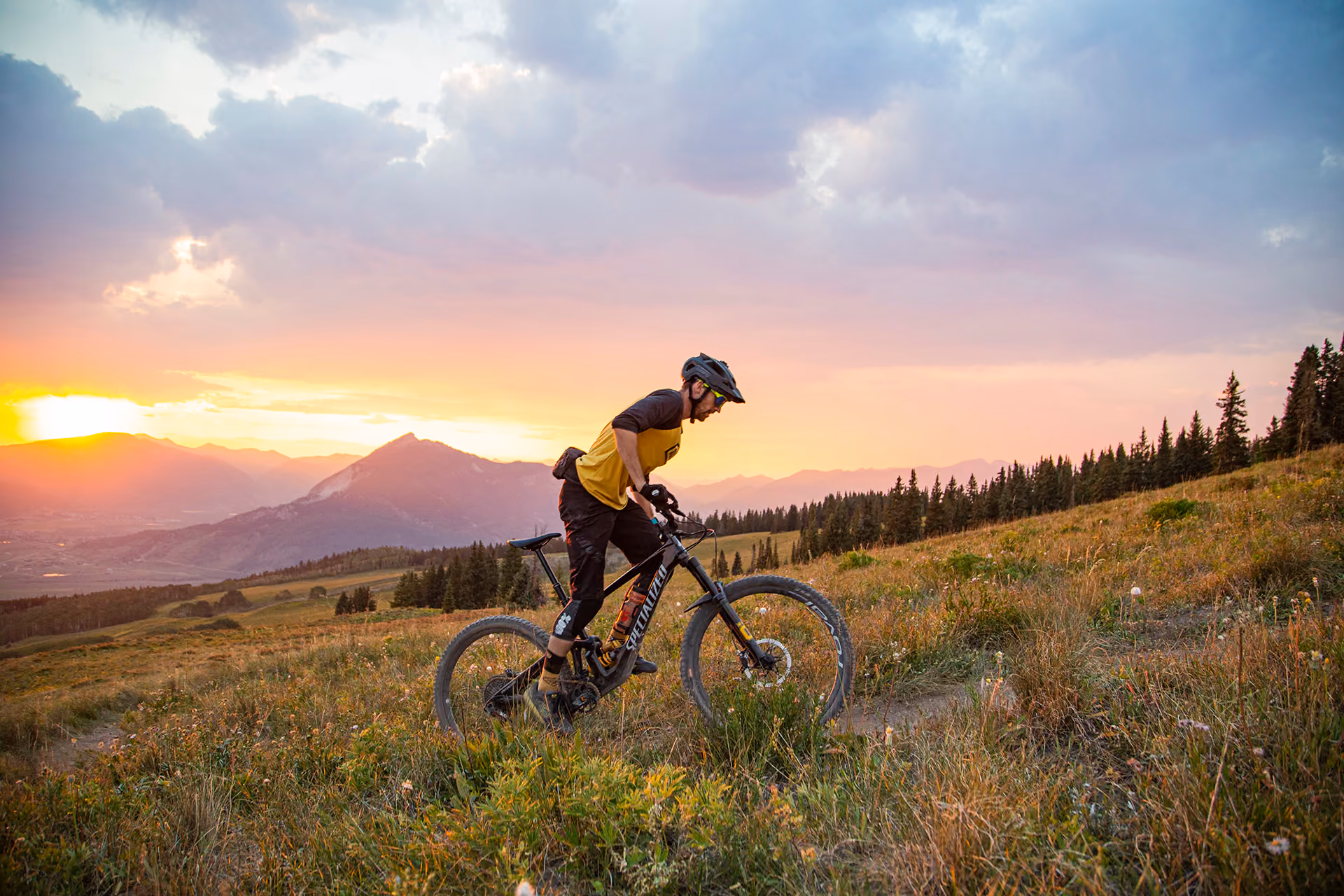 Mountain biker riding uphill on a grassy trail at sunset with mountains and trees in the background.