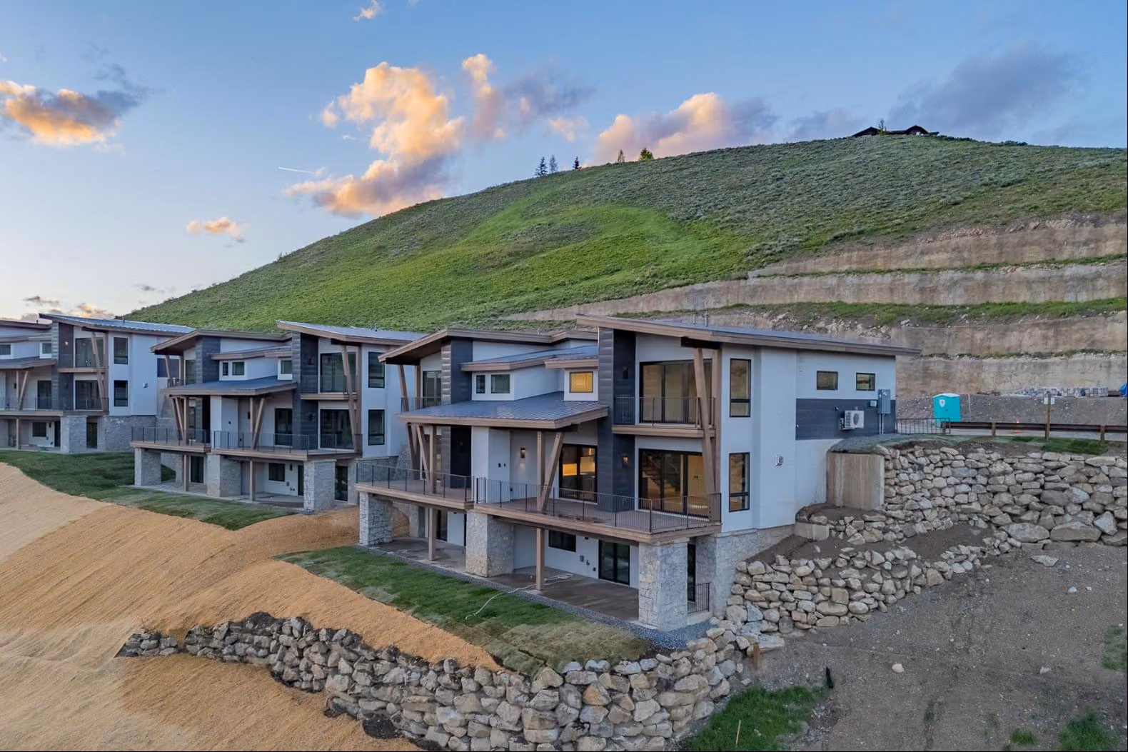 Modern multi-story homes with rock retaining walls on a hillside under a blue sky with clouds.