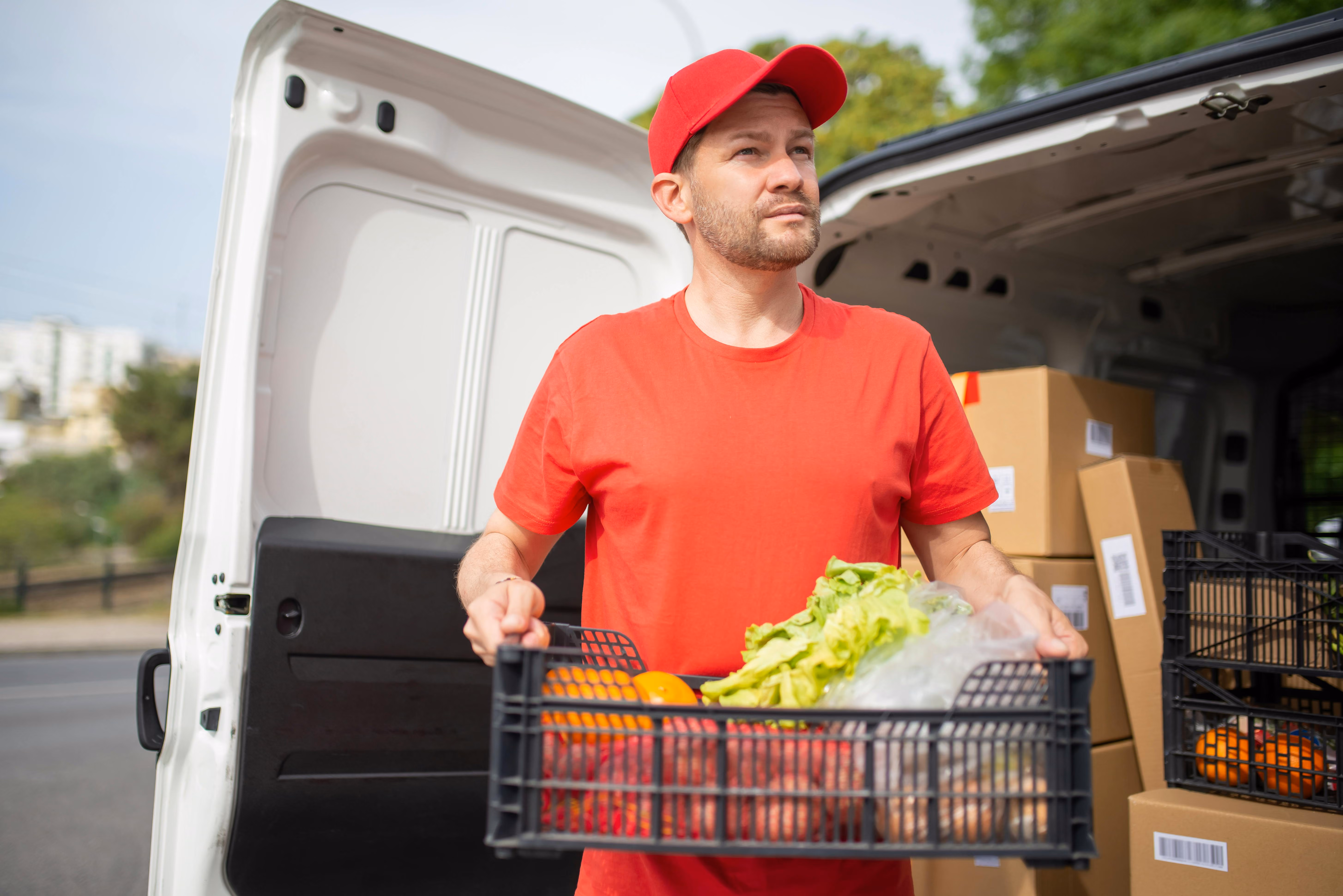 Man in red shirt and cap holding crate of fresh vegetables and fruit by a white delivery van.
