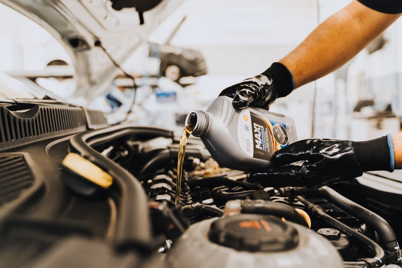 Gloved hands pouring motor oil from a container into a car engine under the hood.