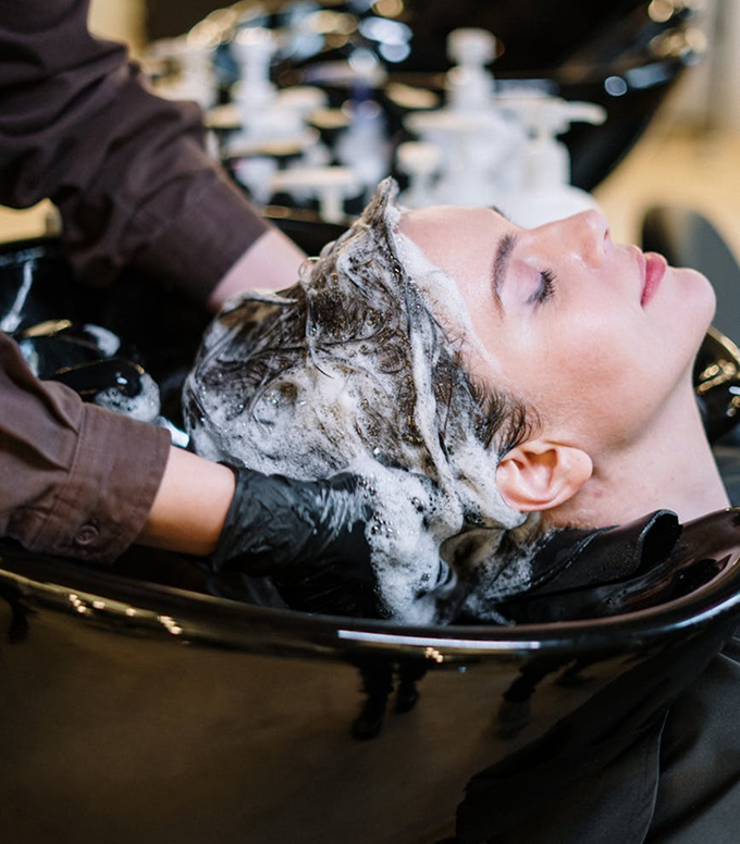 Person with eyes closed getting hair washed with soapy lather at a salon basin.
