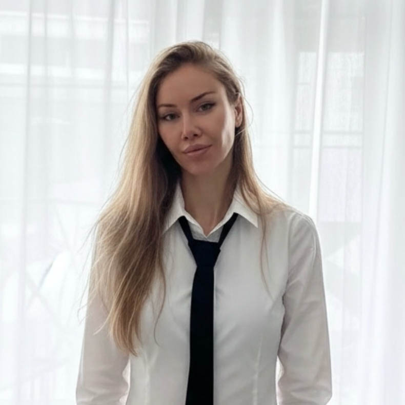 Sarah, who has long blonde hair wearing a white shirt and black tie standing in front of a sheer curtain.